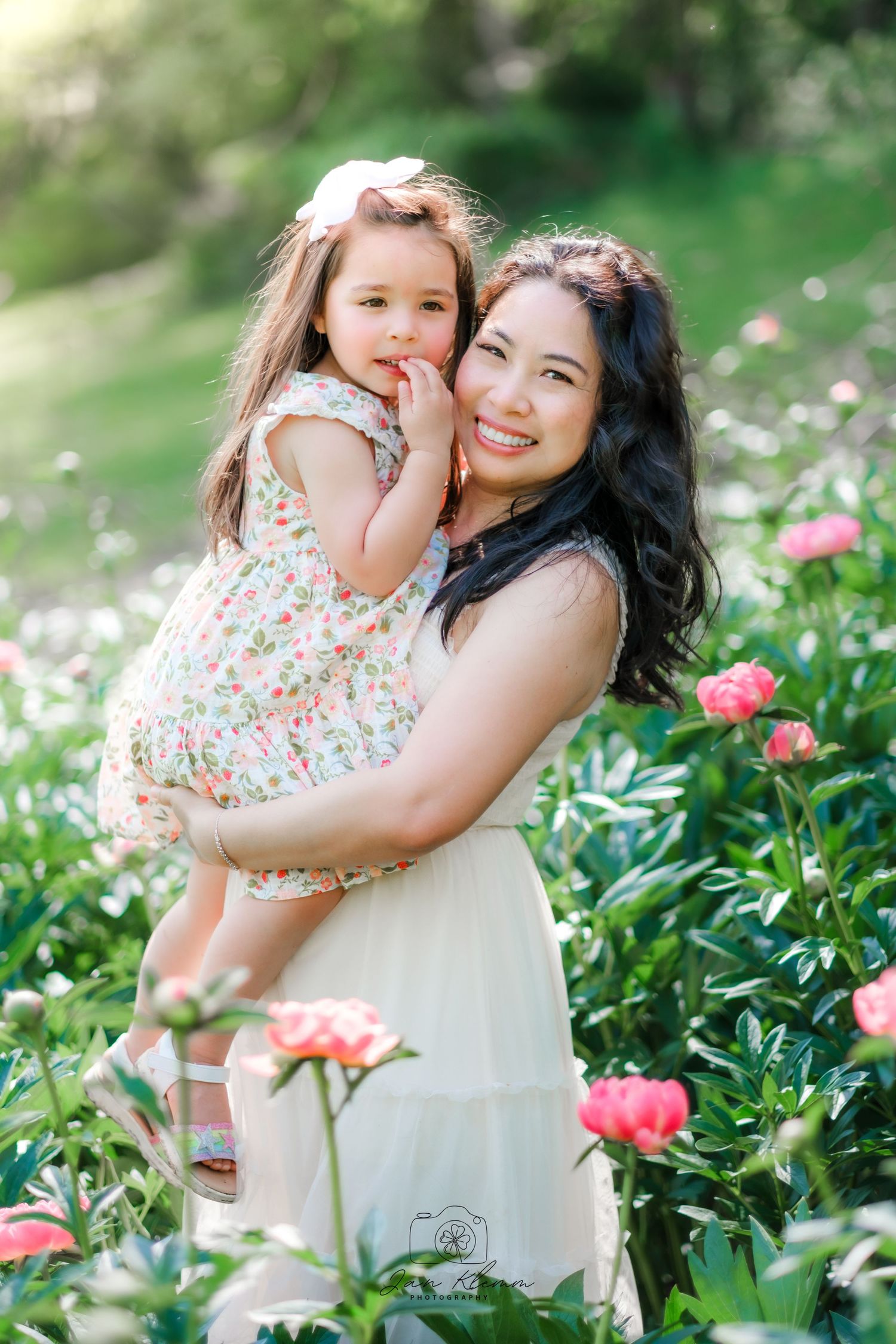 A smiling mother holds her young daughter in a floral dress among pink peonies in a lush garden.