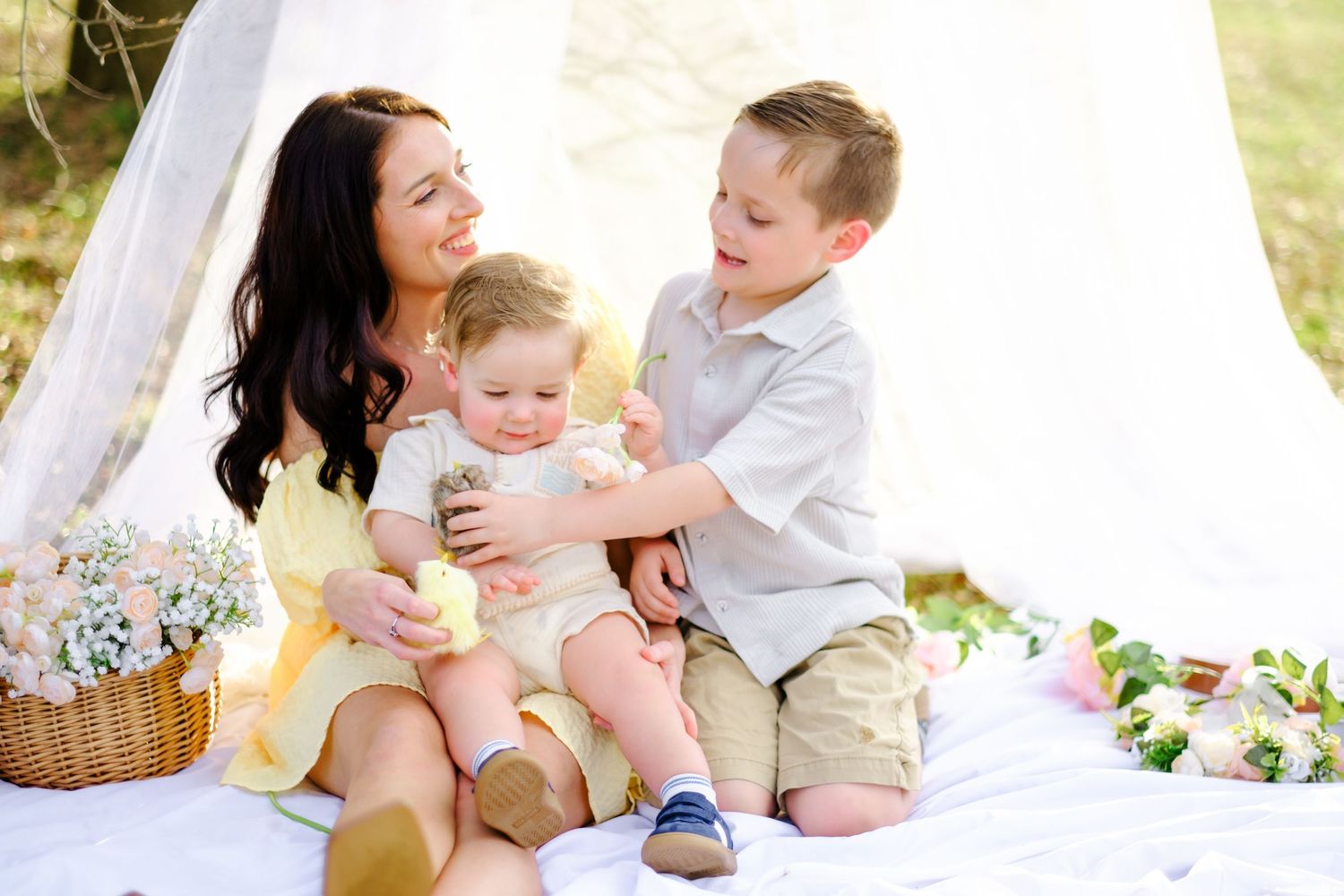 Mother in yellow dress sits outdoors with two young sons near white teepee during sunny family picnic session.