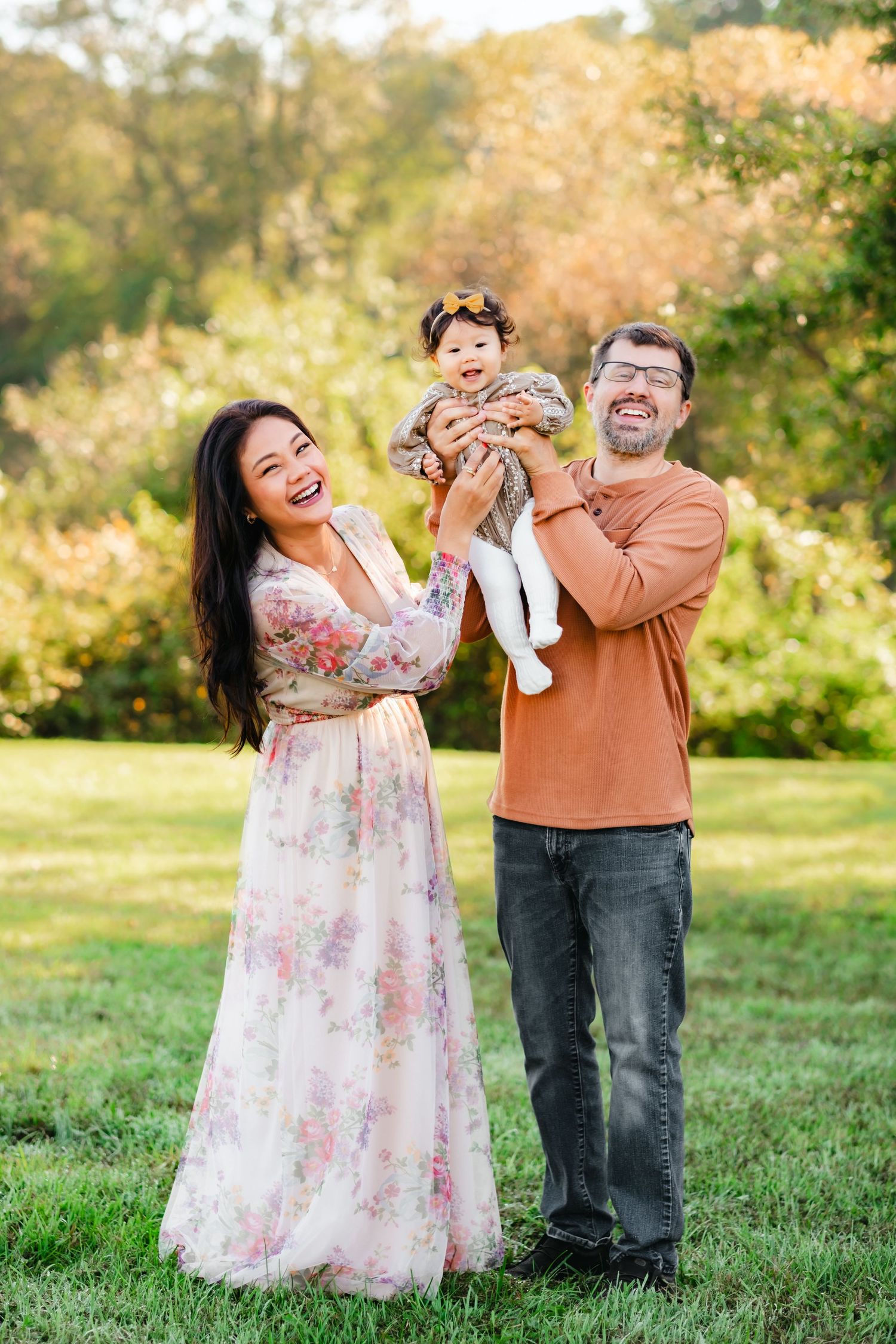 Happy family with mother in floral dress and father in orange shirt lifting smiling baby girl outdoors in autumn park.