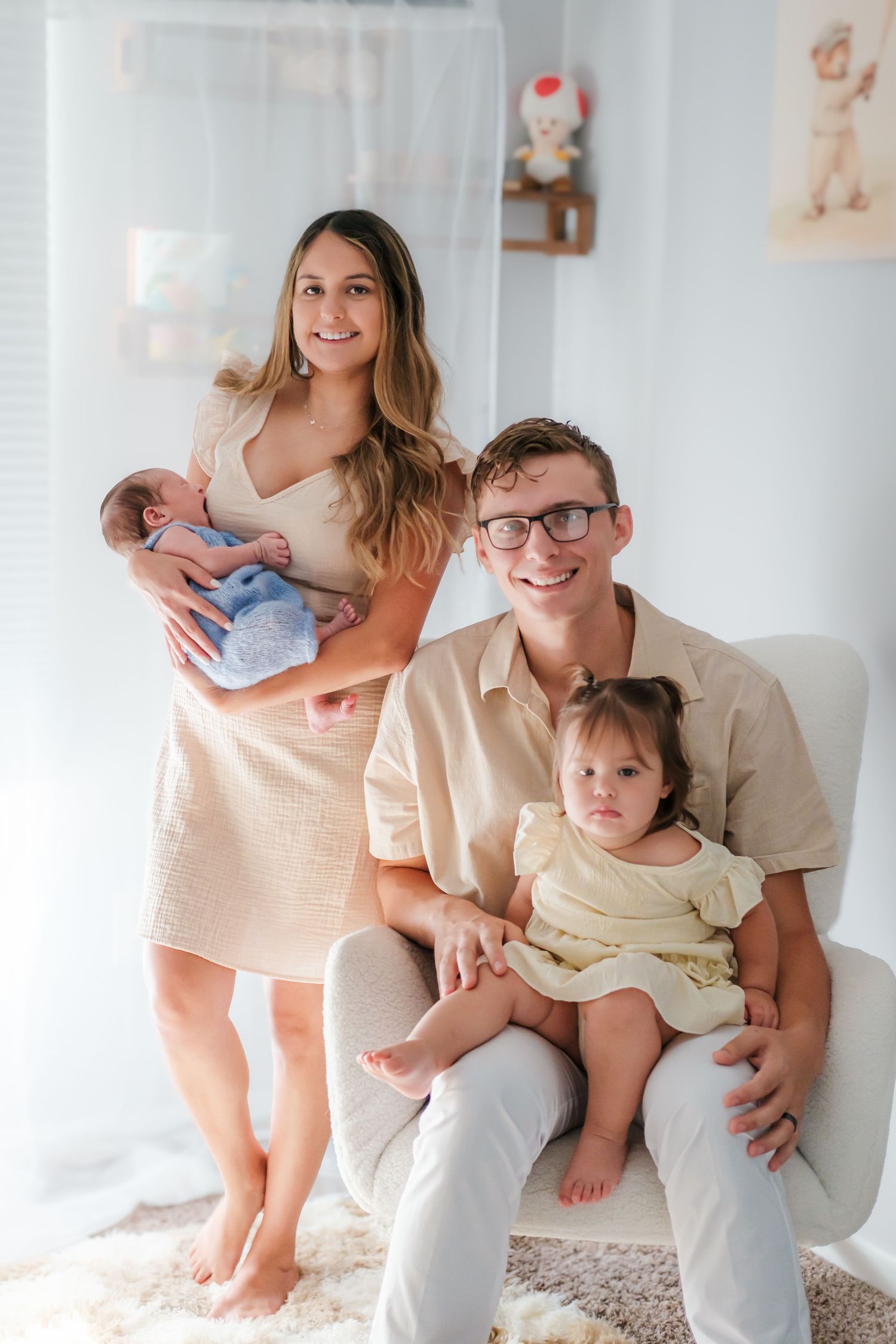 Young family portrait with mom holding newborn, dad seated with toddler daughter, in bright nursery room.