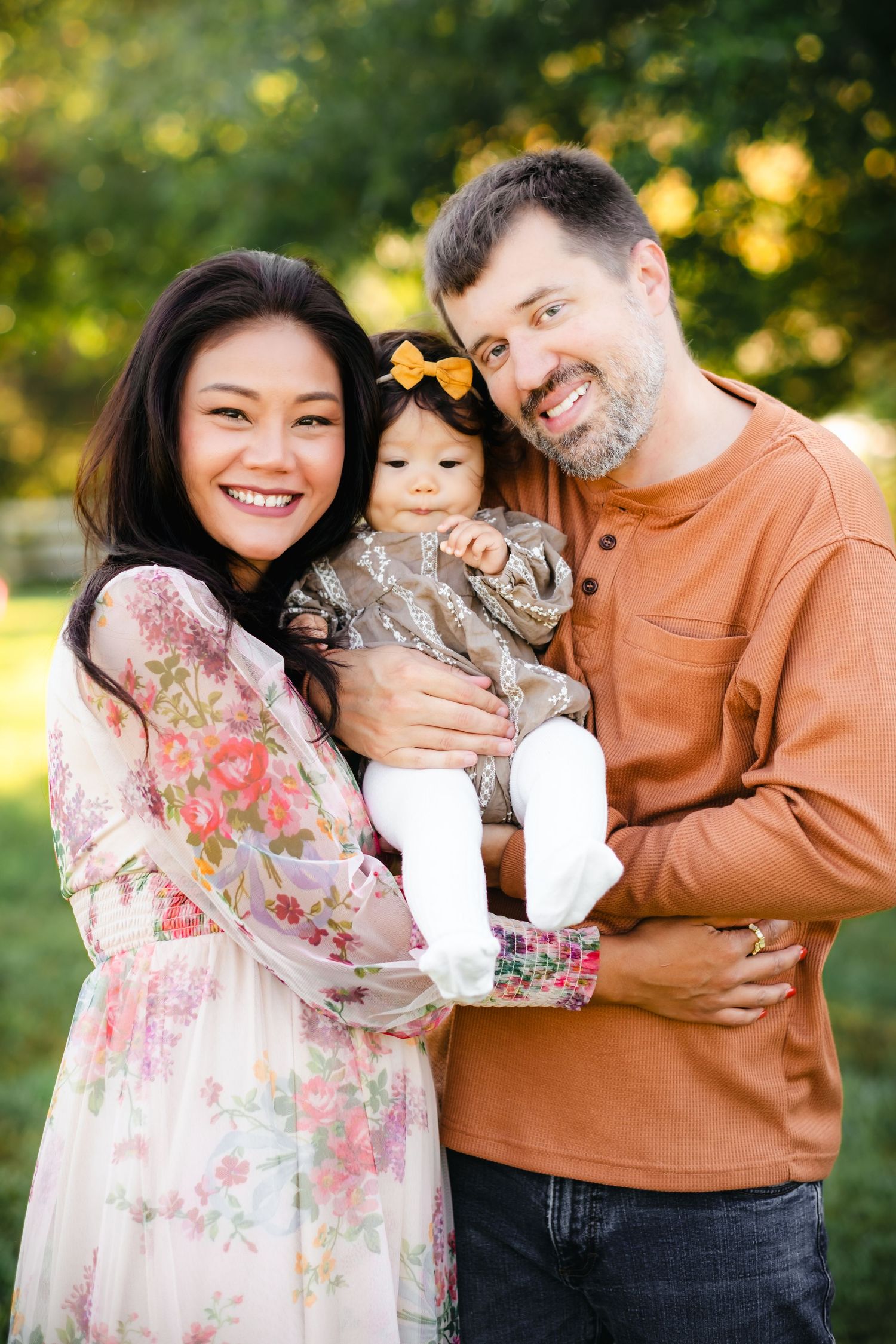Smiling interracial family with mother in floral dress and father in orange shirt holding baby girl with bow outdoors.