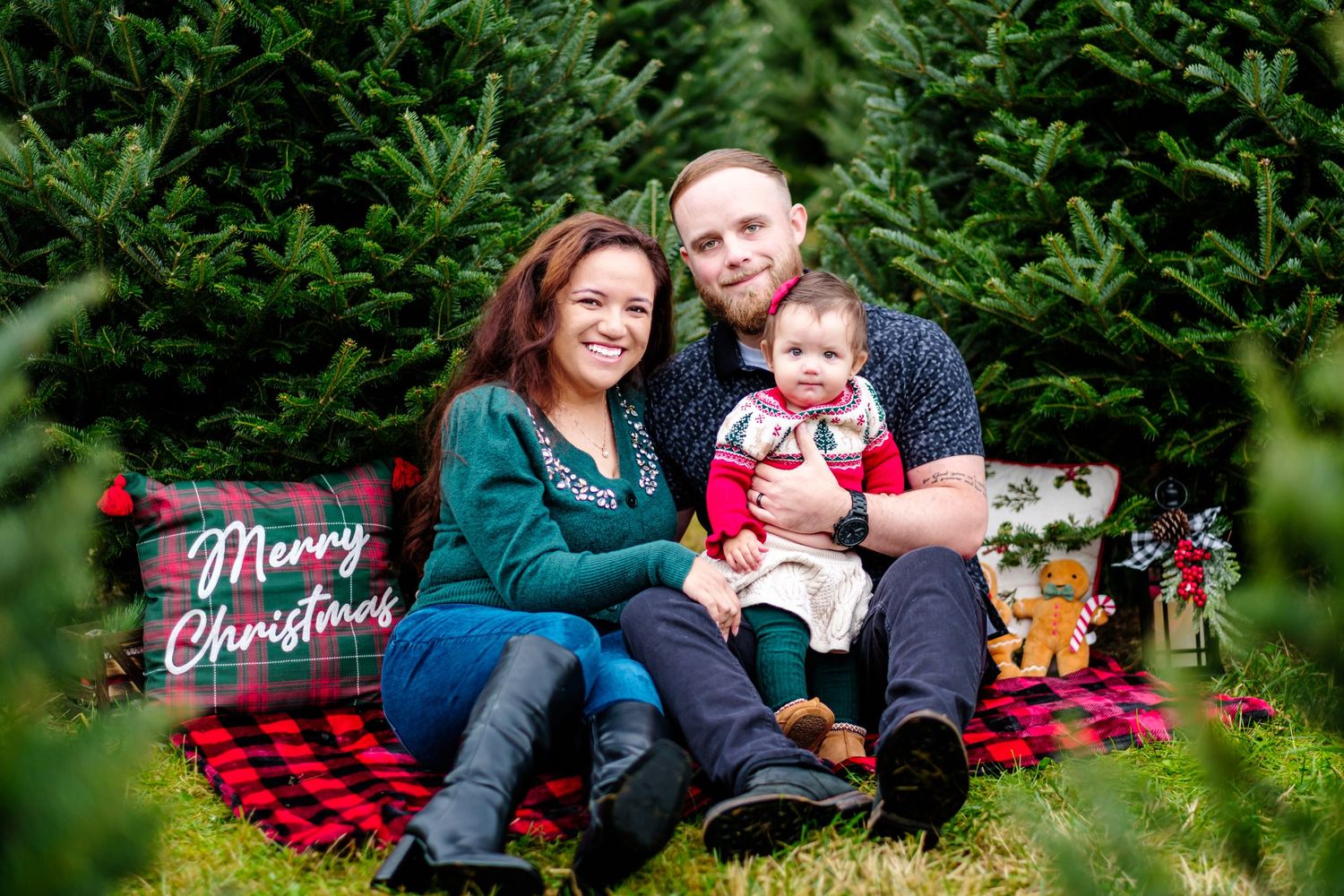 Family of three poses at Christmas tree farm with Merry Christmas pillow, mom in teal, dad in gray, baby in red.
