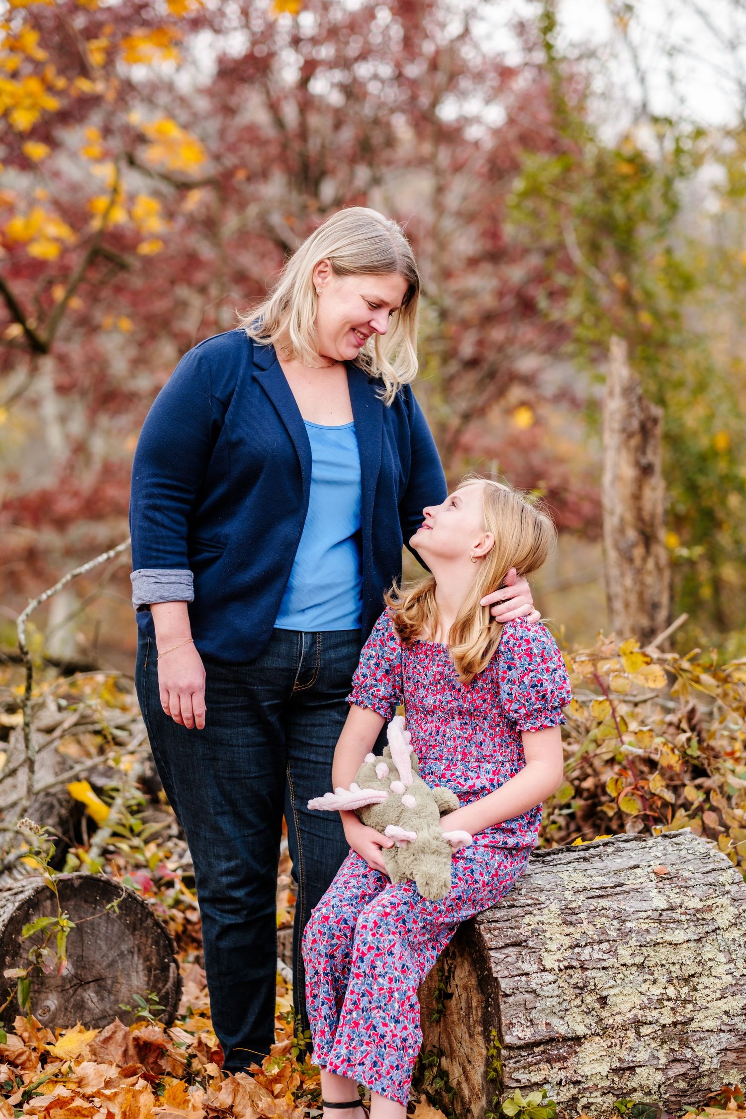 A woman and young girl share a warm moment outdoors among autumn foliage, girl holding a stuffed bunny.