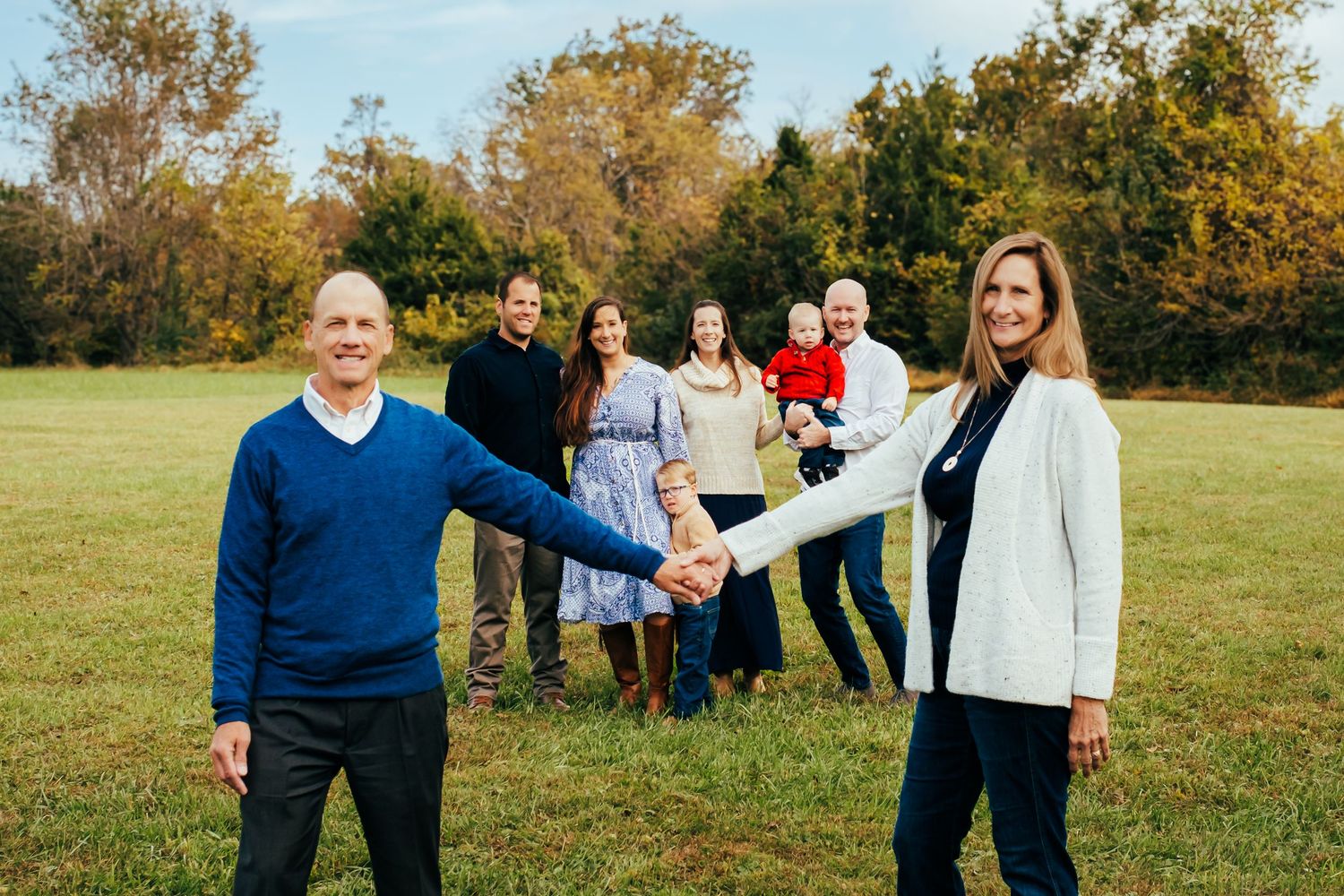 Family of seven poses in a green field, parents holding hands in foreground with adult children and grandchild behind them.