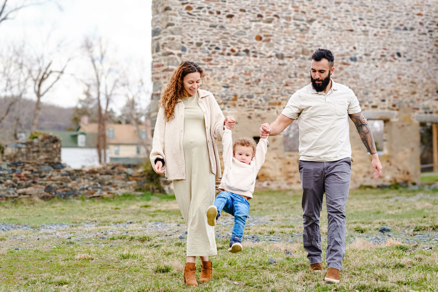 Pregnant woman and bearded man hold toddler's hands while walking outdoors near a stone building.