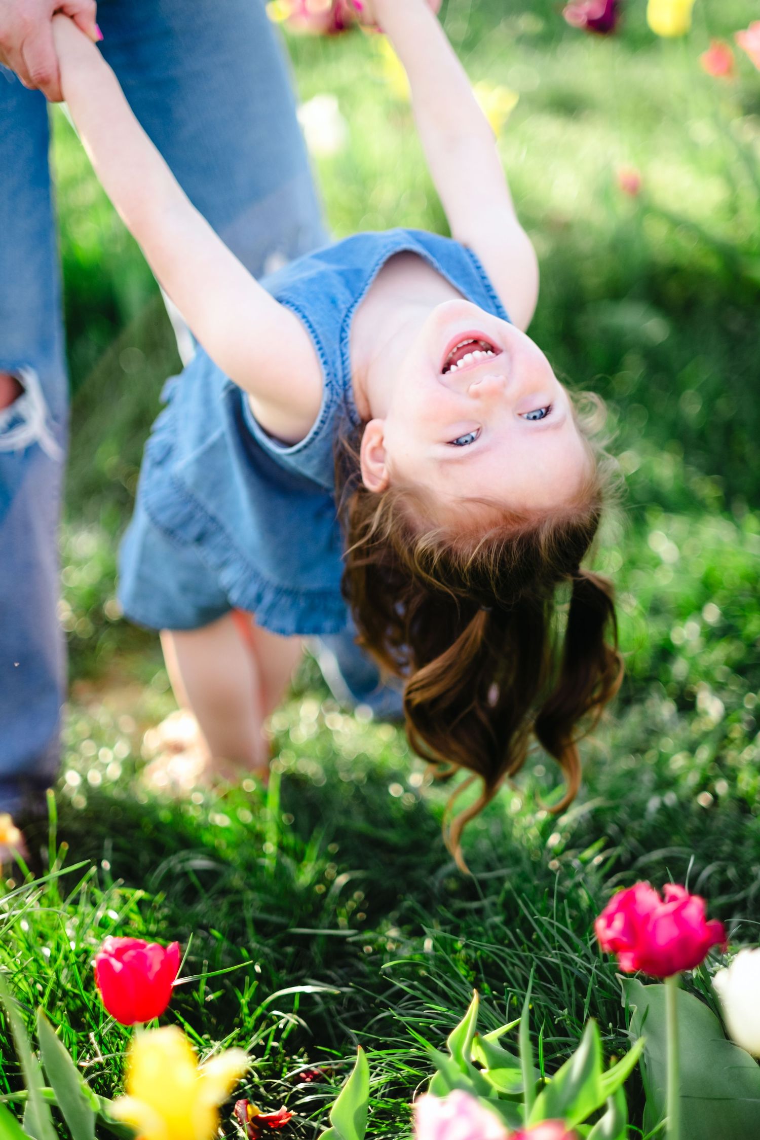A joyful little girl in a denim dress hangs upside down laughing near colorful tulips in a sunny garden.
