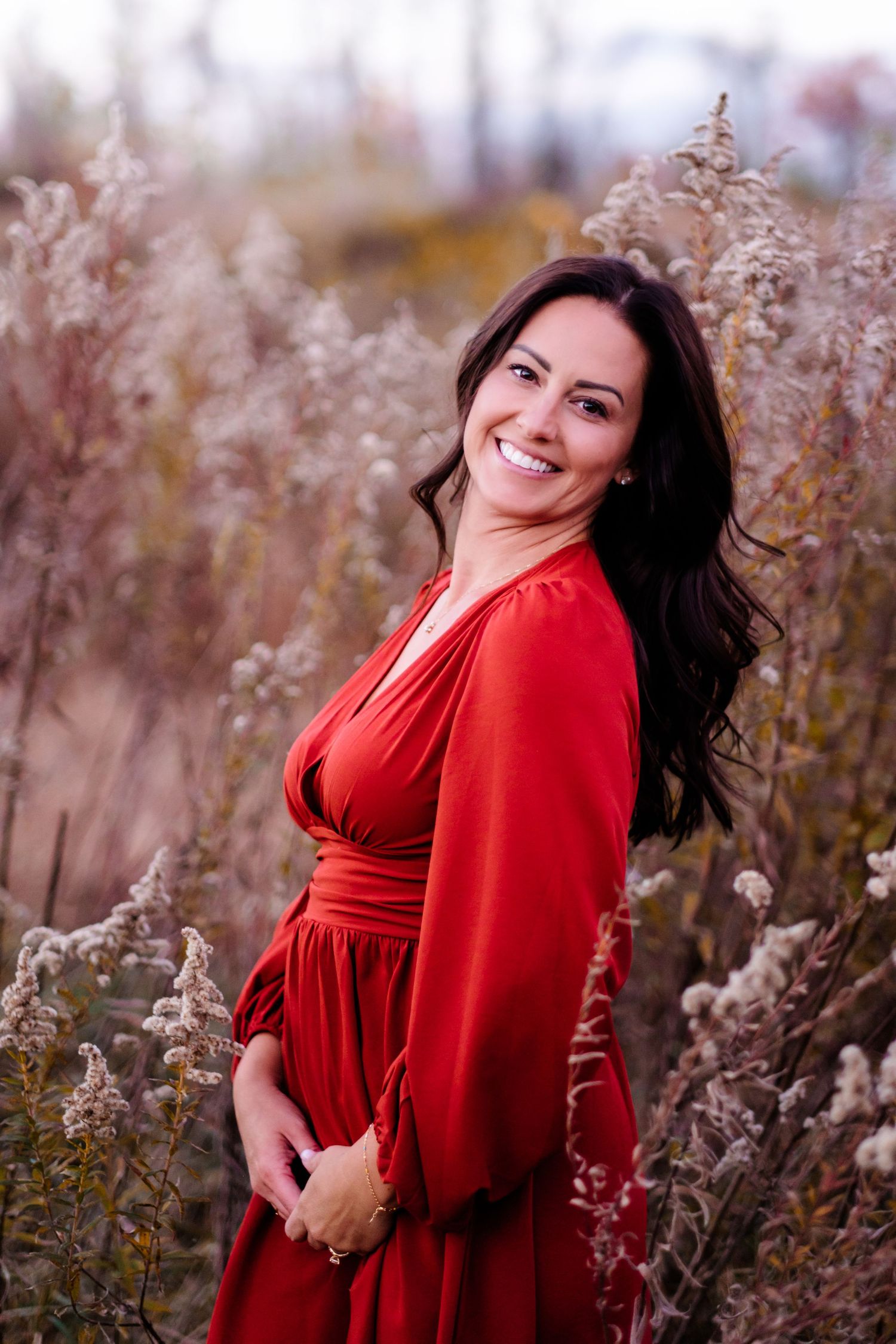 Smiling woman in red dress standing among tall dried grasses in an autumn field.