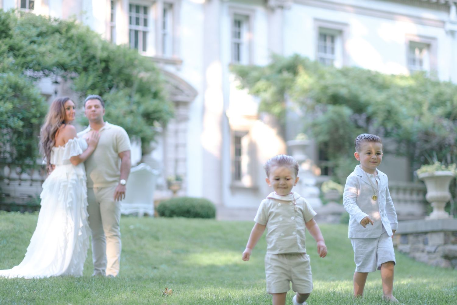 Elegant outdoor wedding scene with couple in white and beige attire, two young boys running on lush green lawn before grand estate.