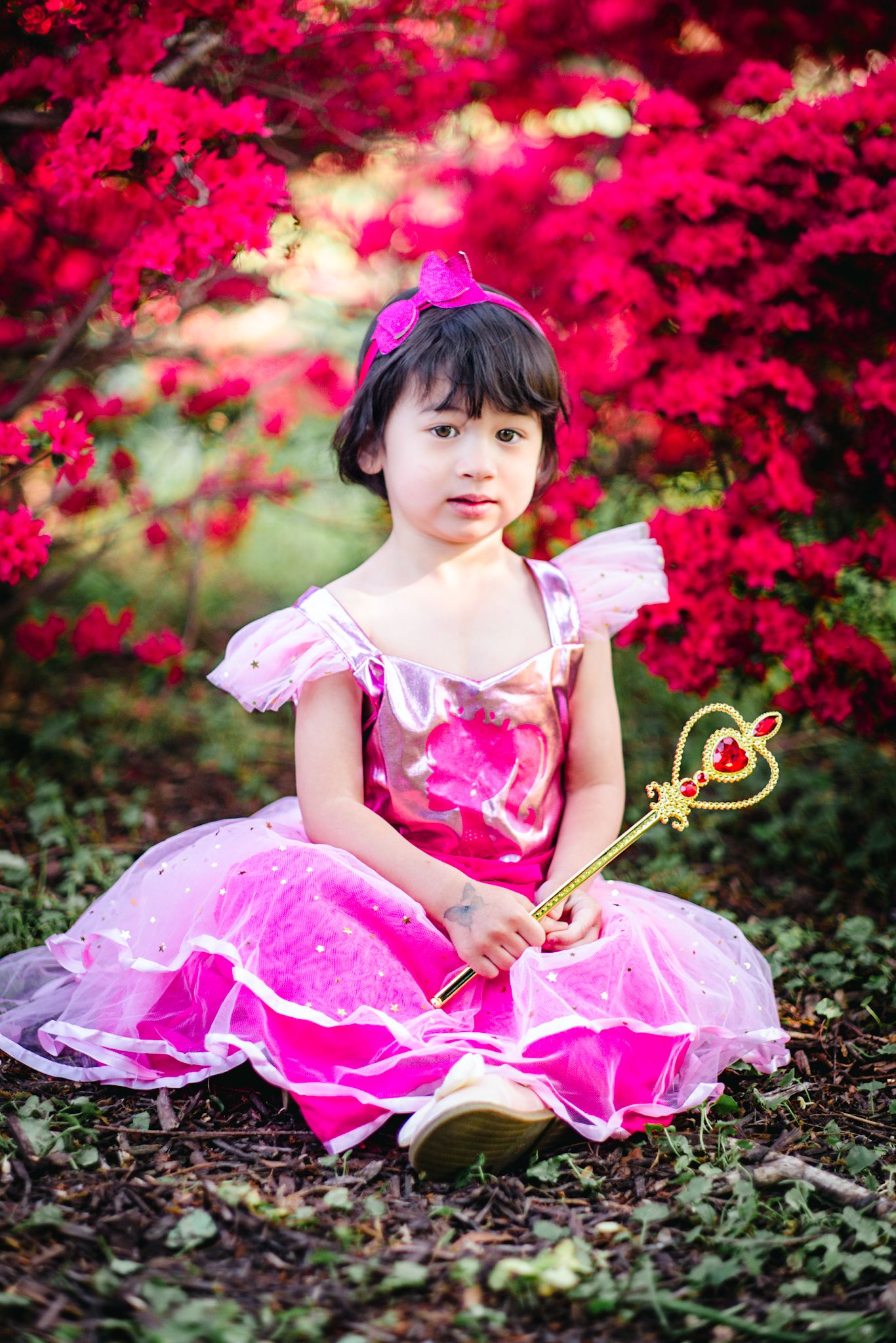 Young girl in pink princess dress holds magic wand, sitting among vibrant red azalea bushes outdoors.