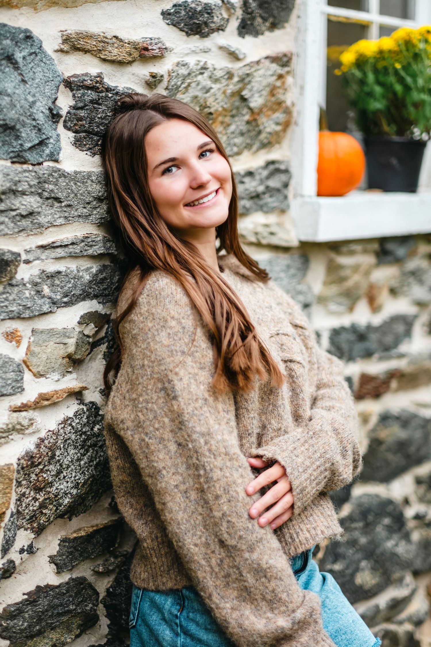 Young woman in beige sweater smiling against stone wall with fall pumpkin decor in background.