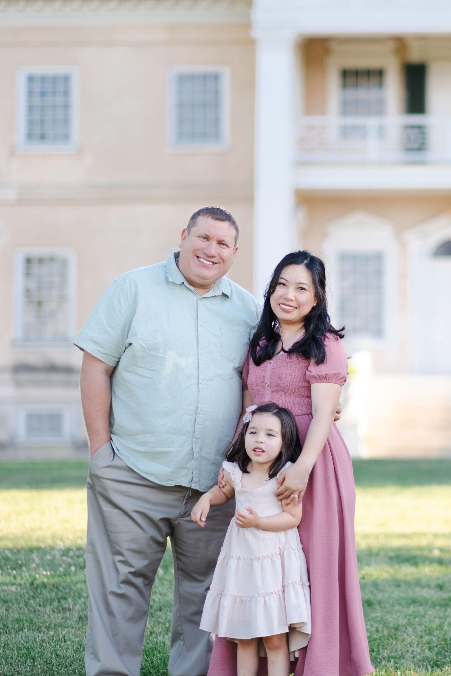 A smiling family of three poses outdoors, with a man, woman in pink dress, and young girl in front of a historic mansion.