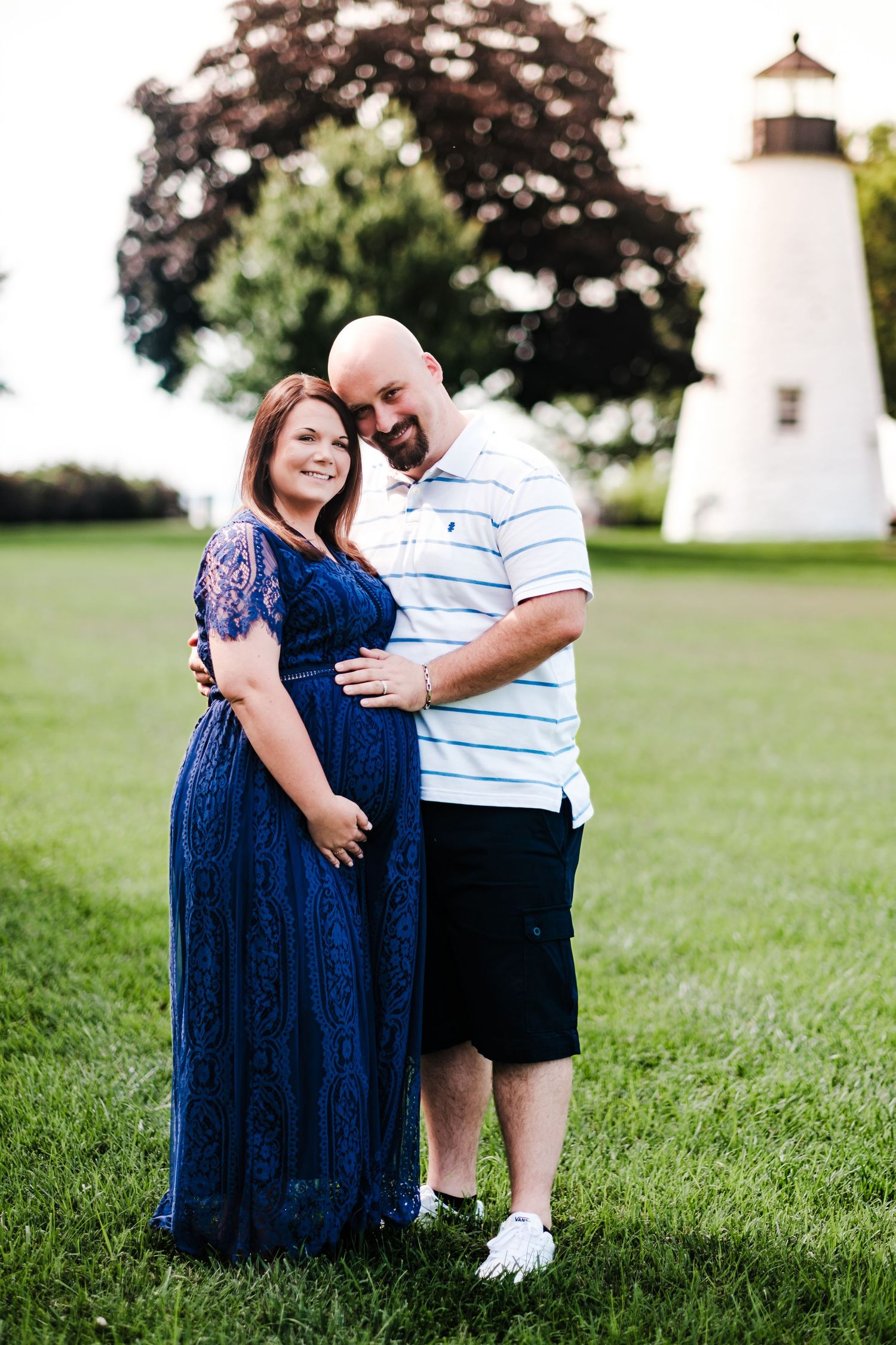 Pregnant couple posing together on green lawn with lighthouse in background during maternity photo session.