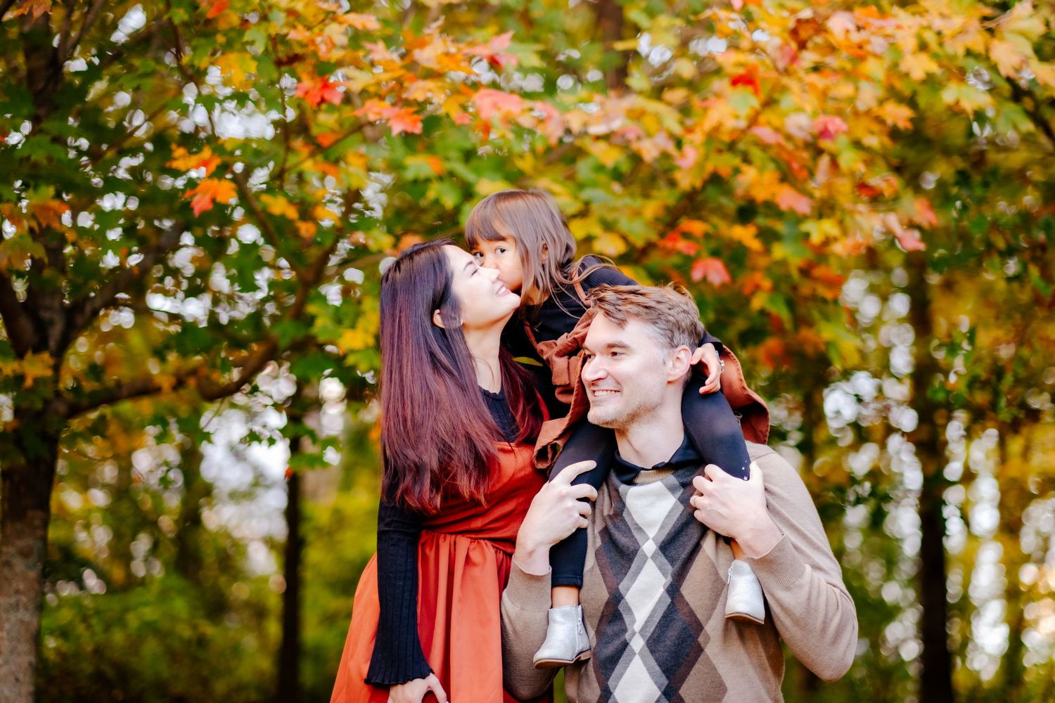Family enjoying autumn park, woman in red dress kissing man\u2019s cheek as child sits on his shoulders.
