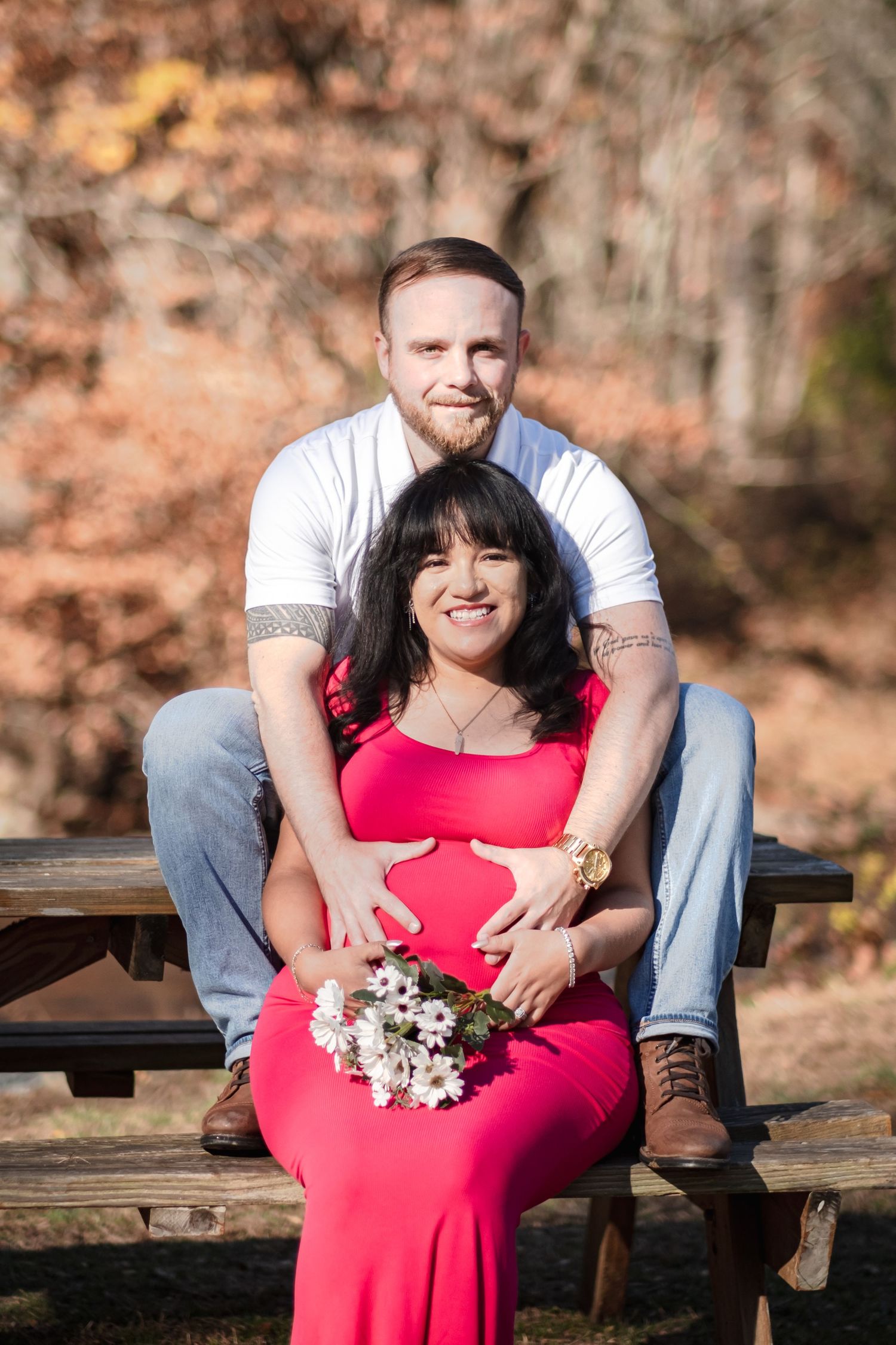 Couple sits on wooden bench during outdoor maternity photoshoot with white flowers and red dress.