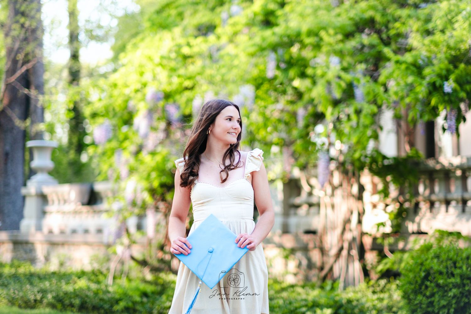Young woman in white dress holding blue clutch purse, smiling outdoors among lush green trees.