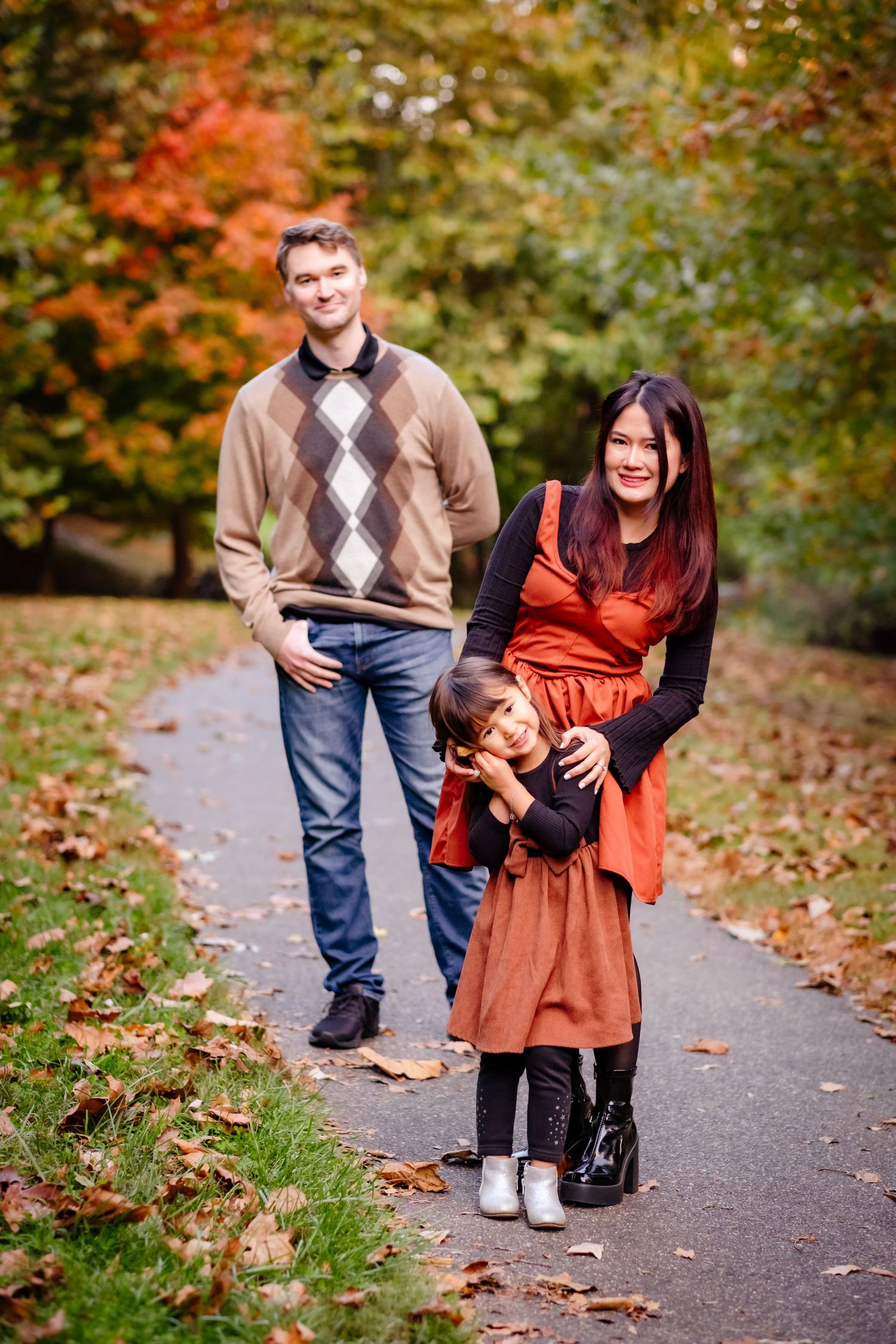 Family of three poses on autumn park path, parents in warm tones with child hugging mom, colorful fall foliage background.