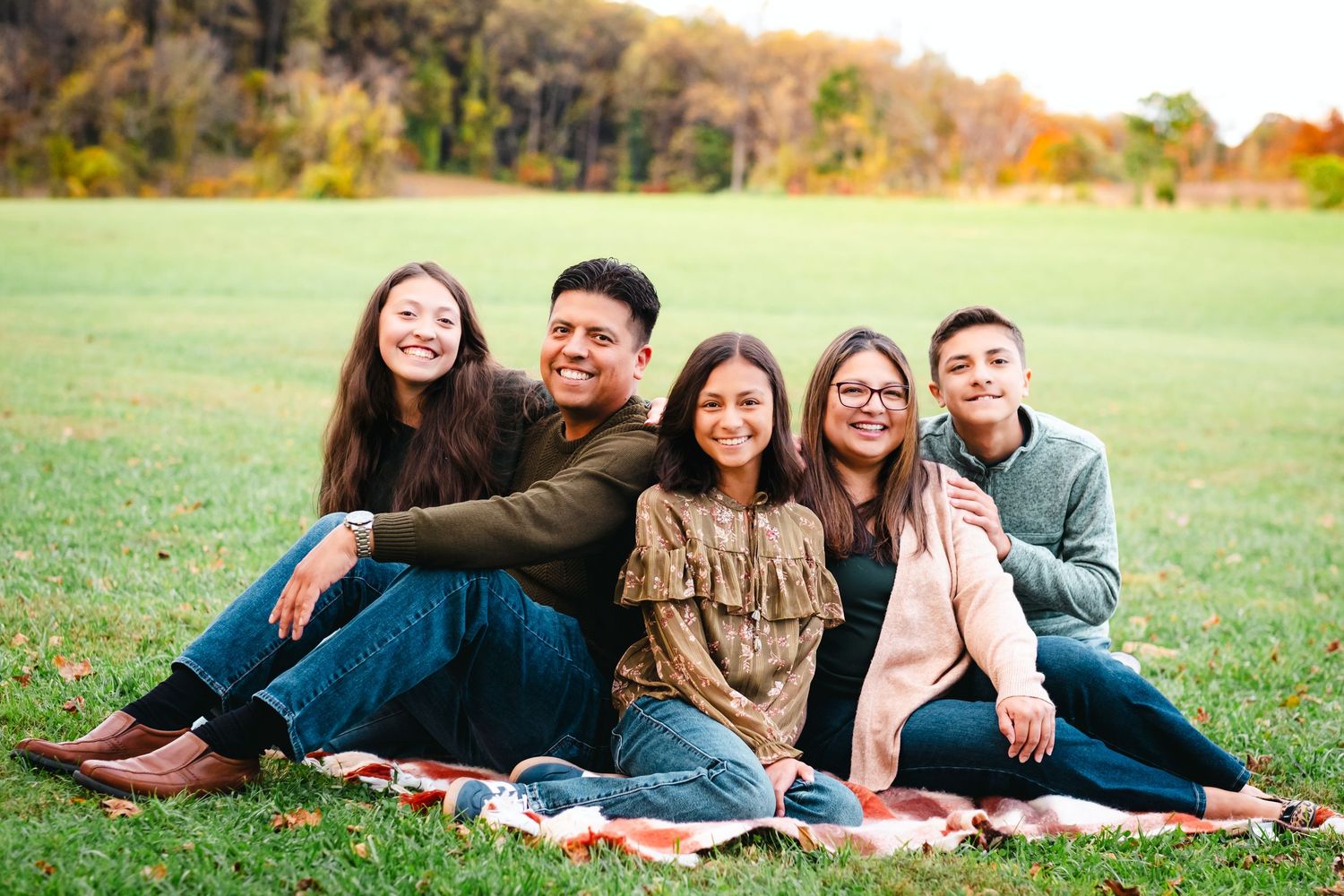Happy family of five sitting together on a blanket in a green park with autumn trees in the background.