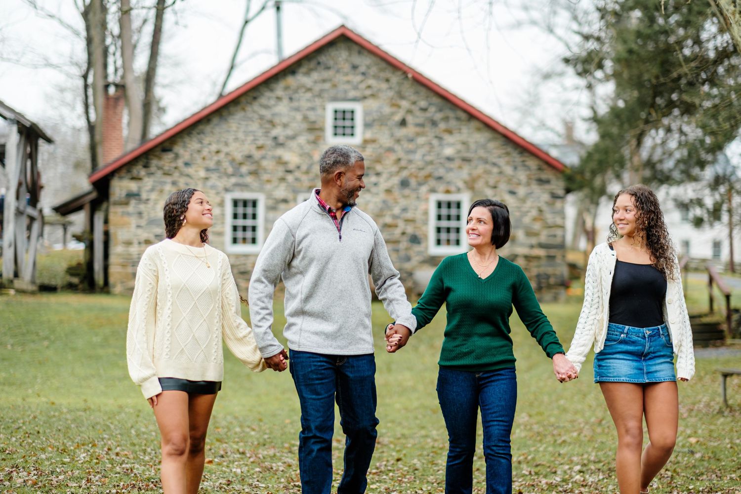 A happy family of four holds hands while walking outdoors near a rustic stone house surrounded by trees.