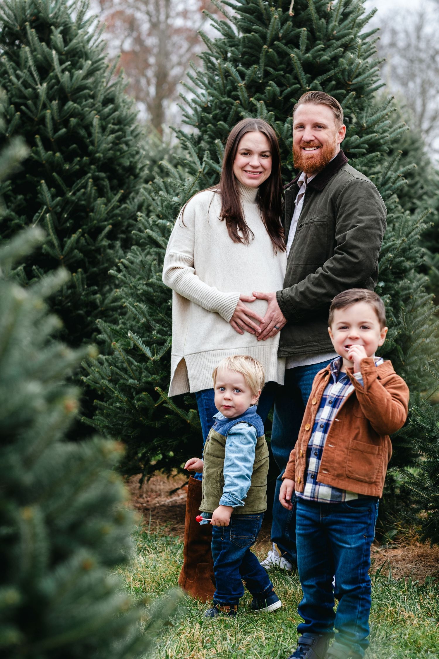 Family of four posing at a Christmas tree farm, parents holding hands with two young boys surrounded by evergreen trees.