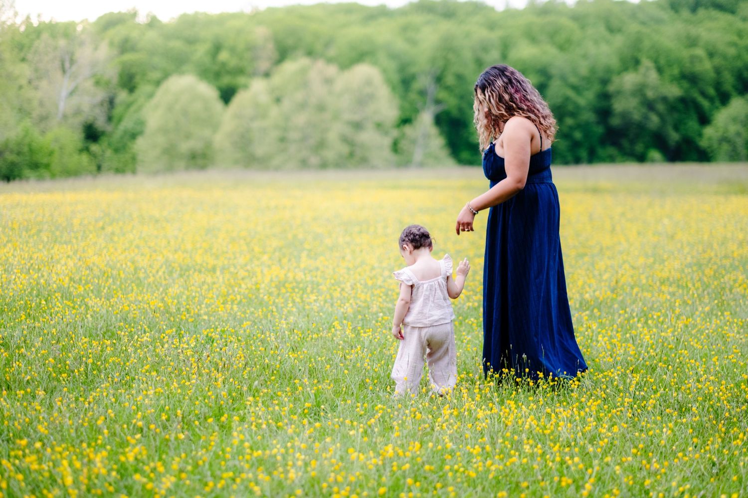 Mother in blue dress walks with toddler through sunny wildflower meadow with green trees in background.