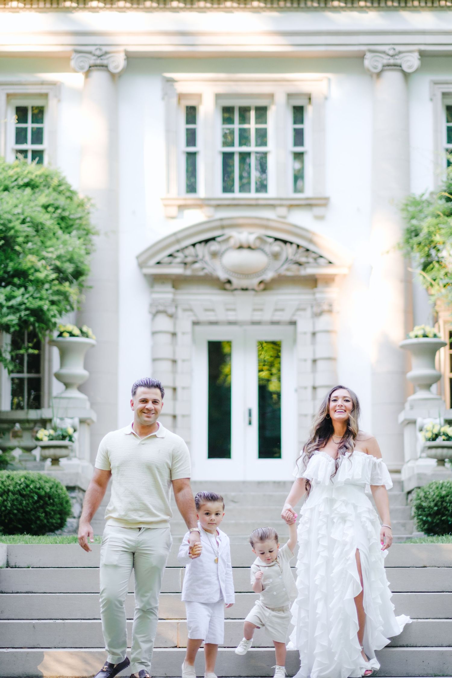 Family of four in white outfits posing on steps of elegant mansion with ornate architecture and lush greenery.