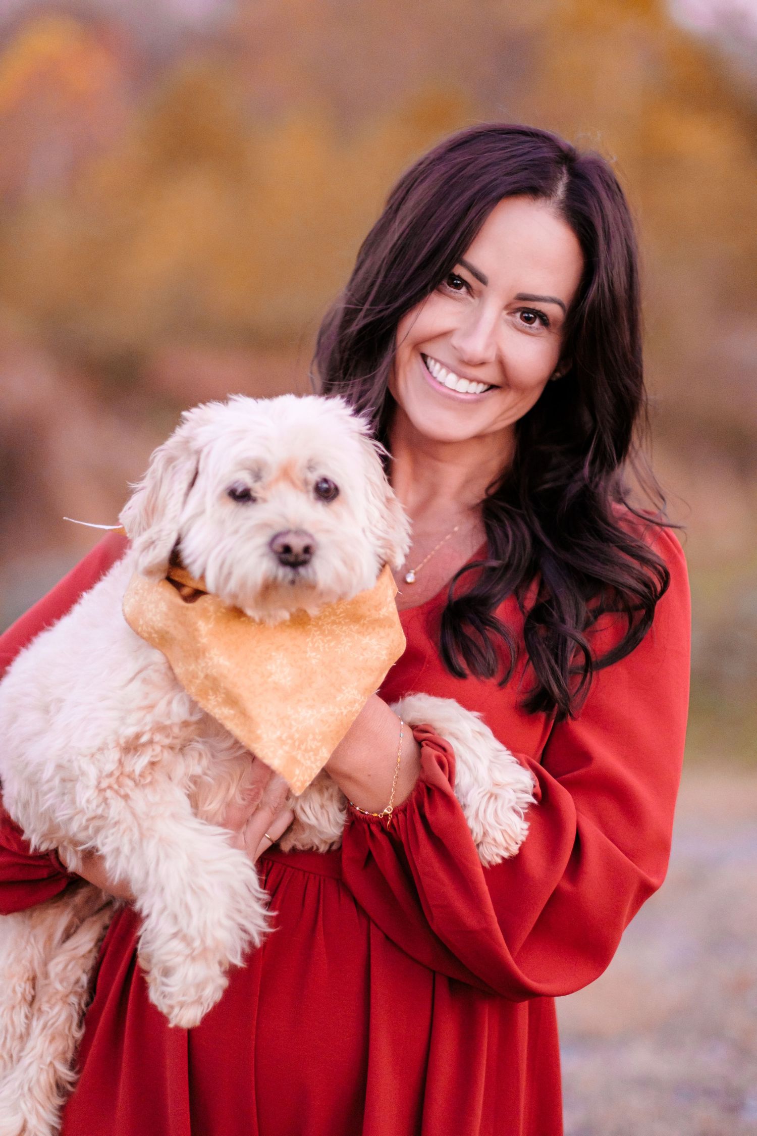 A smiling woman in a red dress holds a fluffy white dog wearing an orange bandana outdoors in fall.