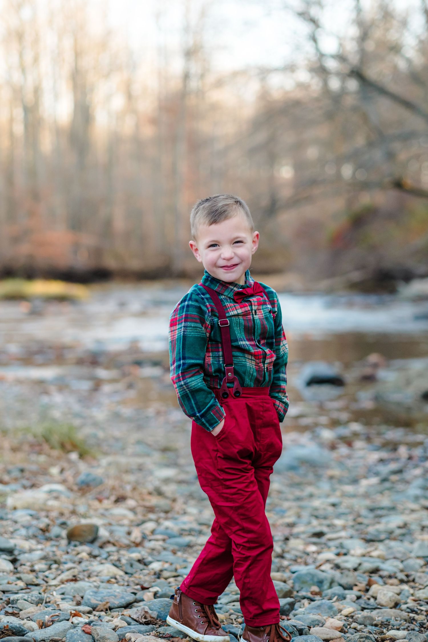 A smiling young boy in red suspenders and plaid shirt poses by a rocky creek in an autumn forest setting.