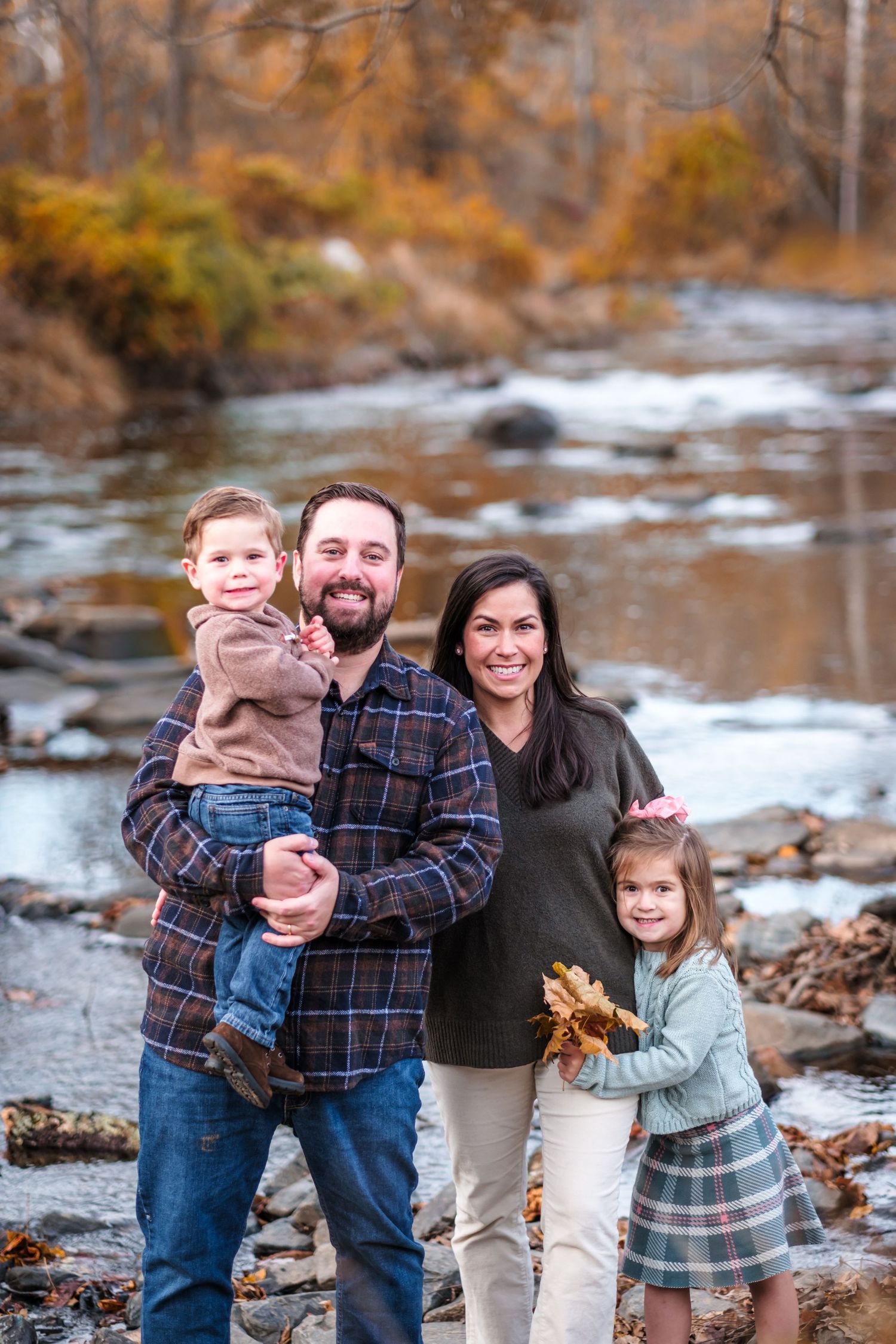 Family of four poses by a rocky stream in autumn, parents holding young children among colorful fall foliage.