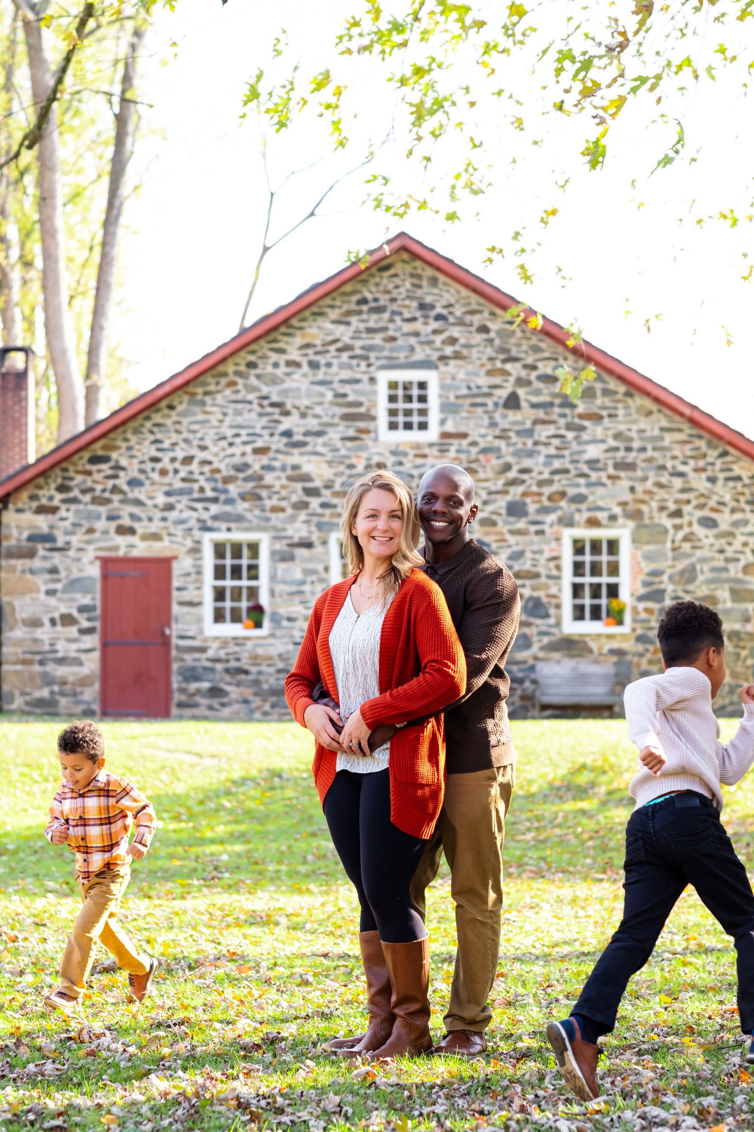 Interracial family posing outdoors in autumn near a stone farmhouse, with two children playing in the background.