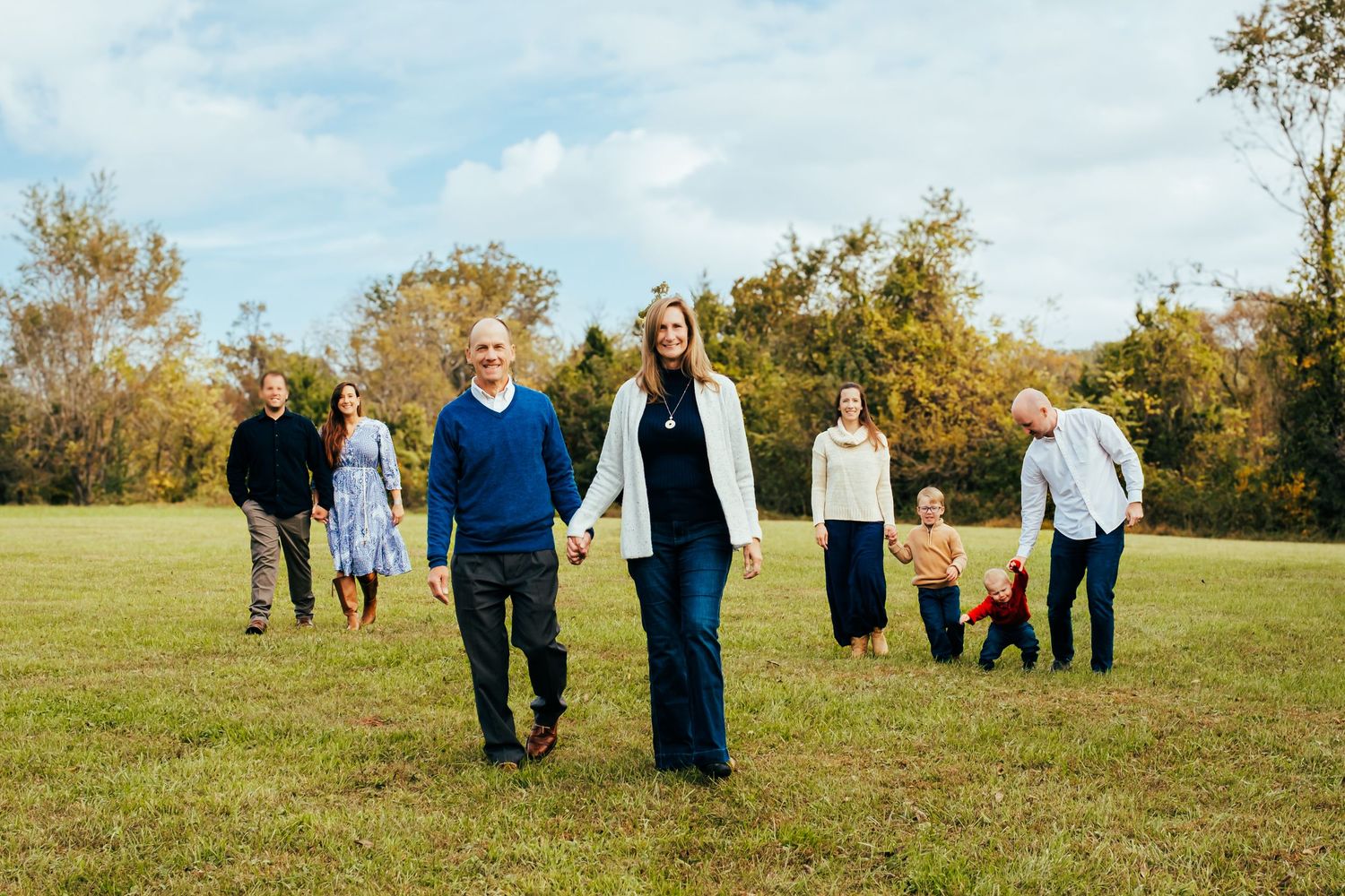 Family group walking in a green field with autumn trees, smiling and holding hands on a sunny day.