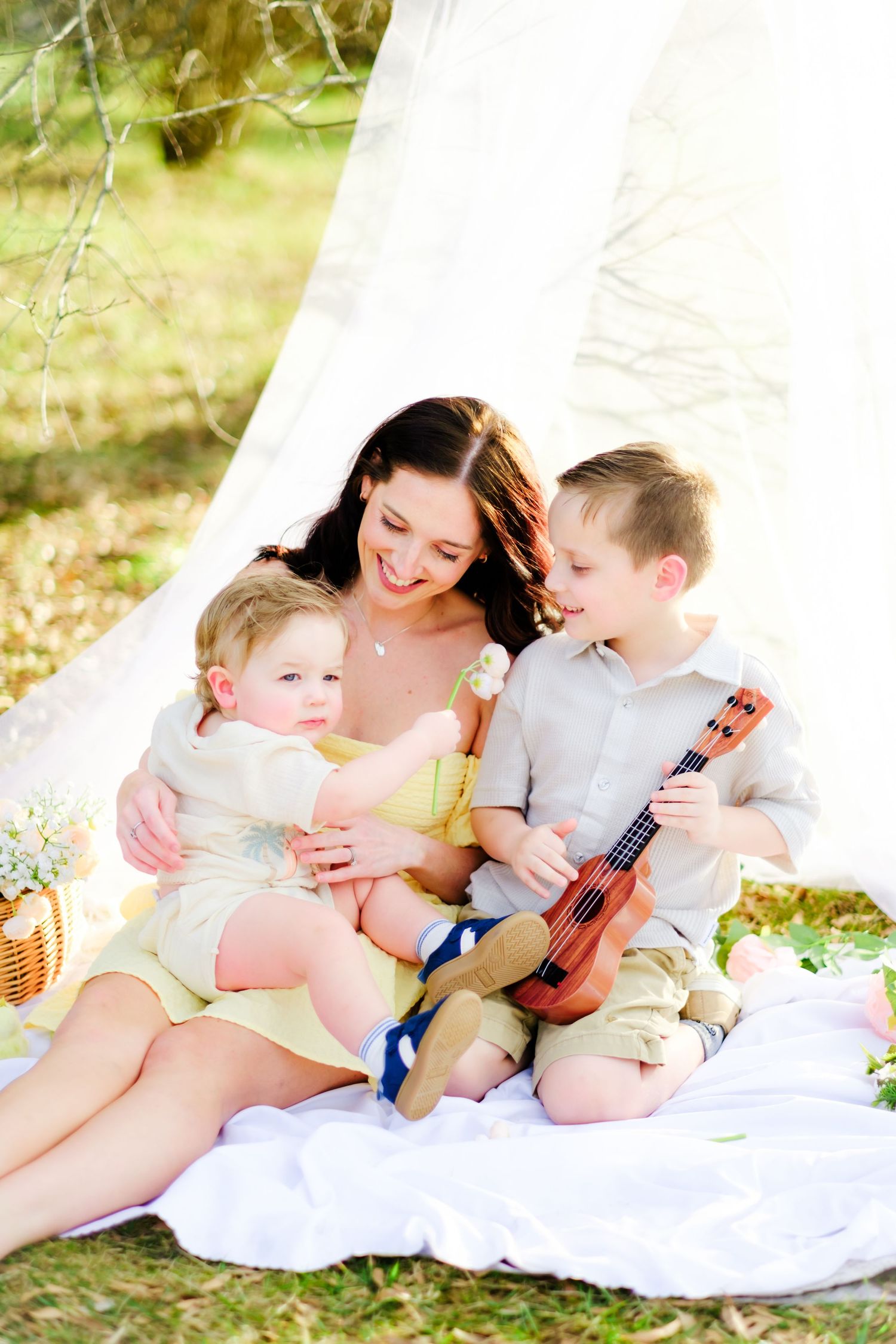 A smiling mother sits with her two young children under a white canopy outdoors, as her son plays a small ukulele.