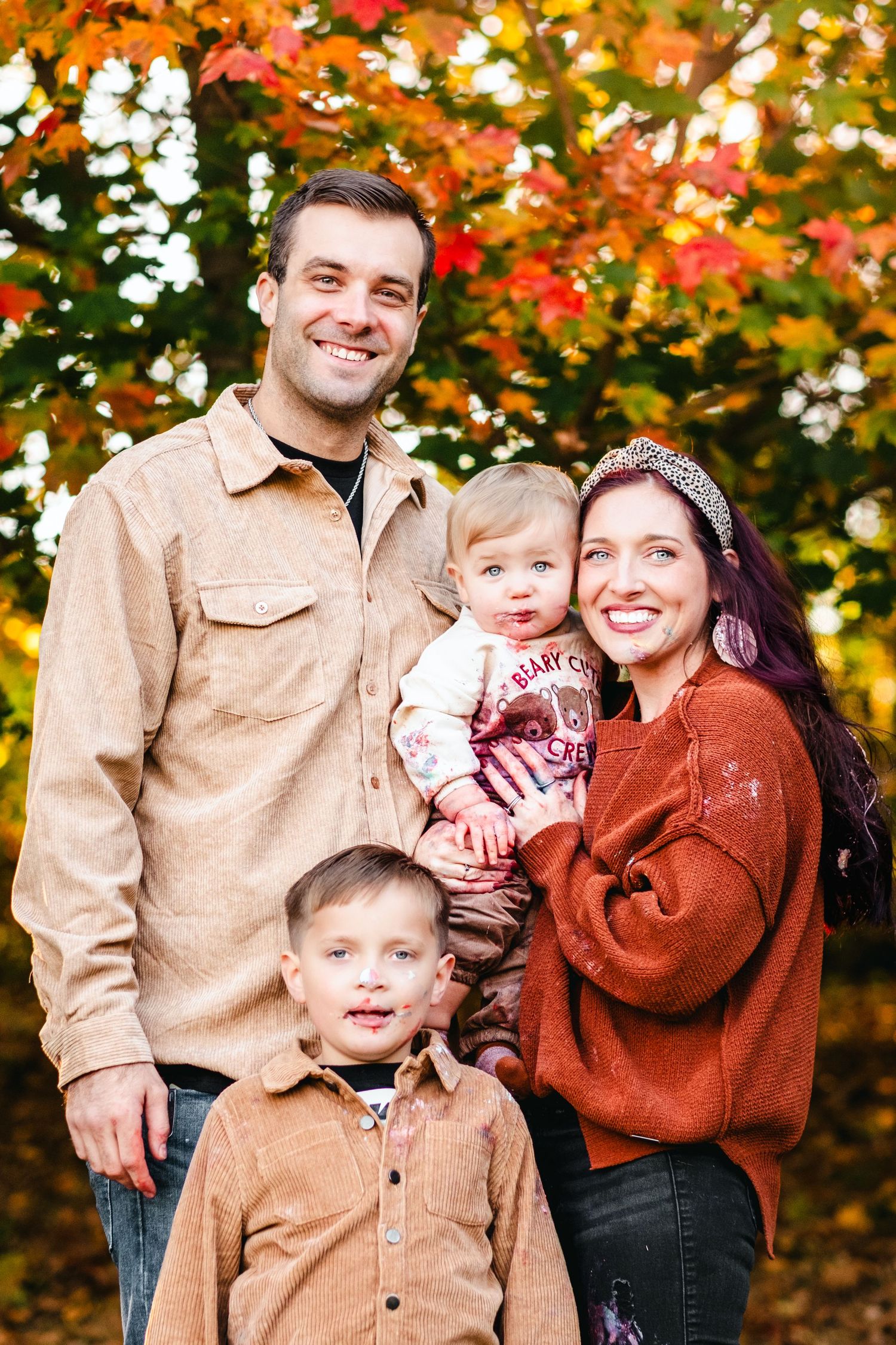 Family of four posing outdoors among vibrant fall foliage, smiling couple holding toddler with young boy in front.