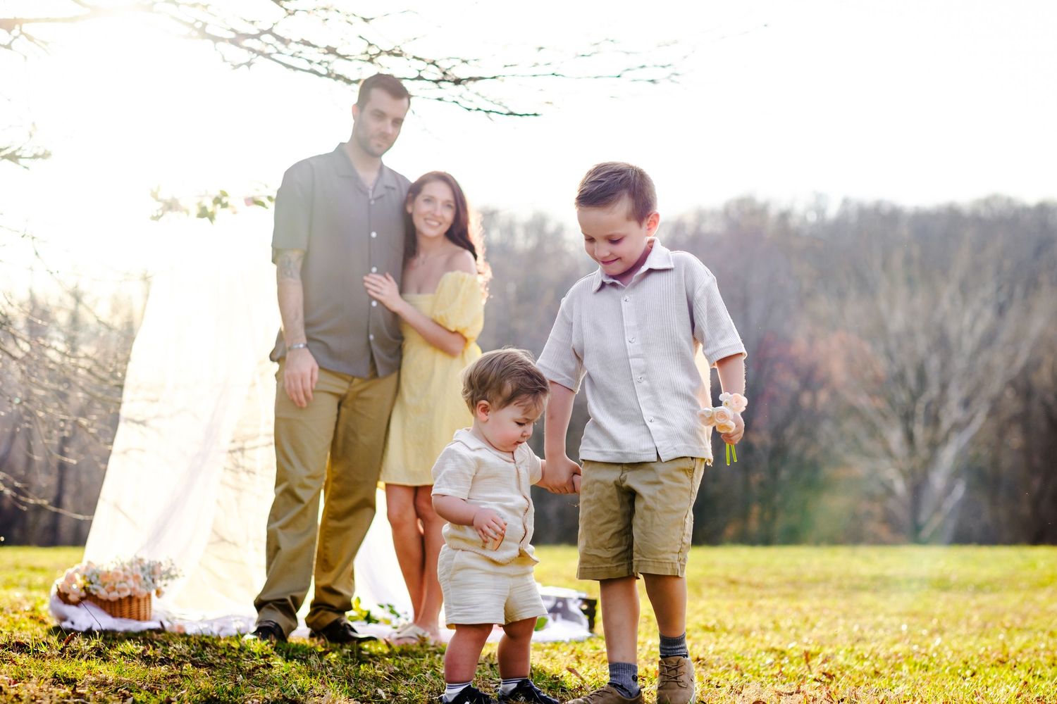 Family of four enjoying a sunny outdoor picnic, with two young boys playing on green grass while parents smile in background.