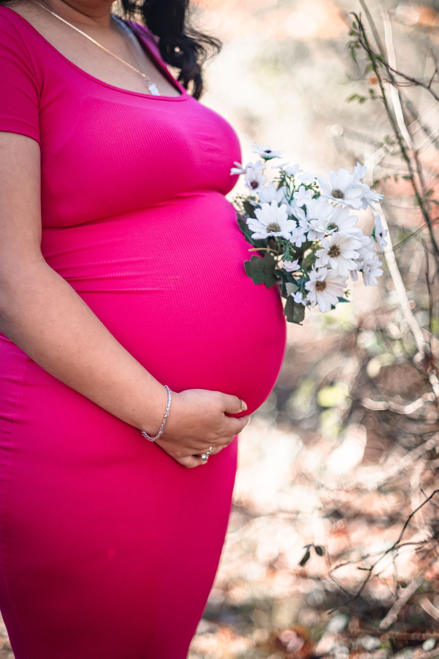 Pregnant woman in pink dress holding white daisy bouquet outdoors, cradling baby bump with bracelet-adorned hand.