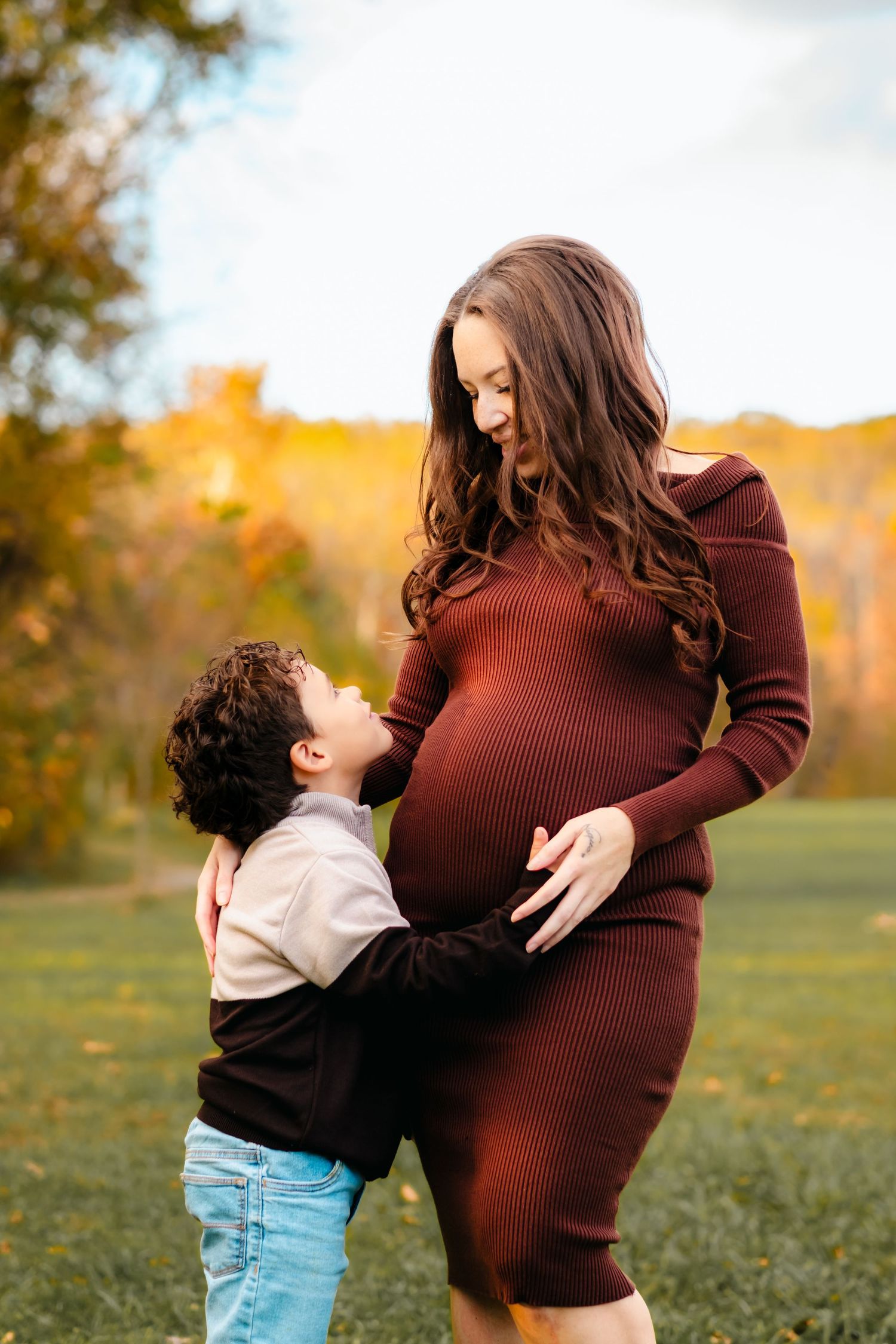 A pregnant woman in a burgundy dress embraces her young son outdoors during a fall maternity photoshoot.