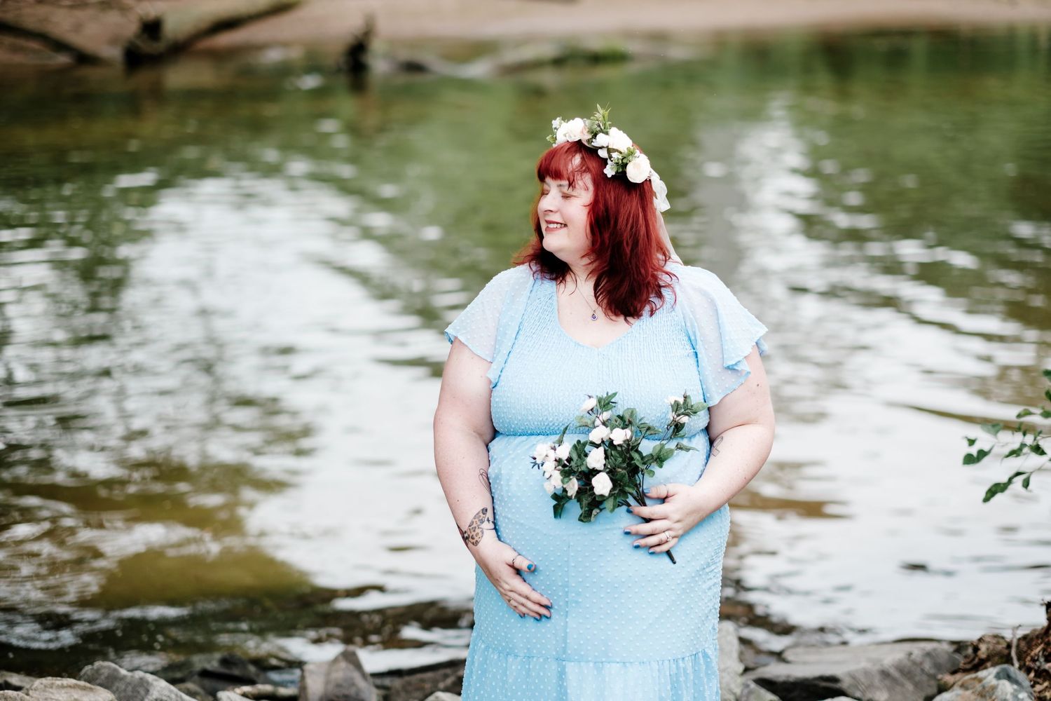 Pregnant woman in light blue dress with floral crown holds bouquet by a serene river.