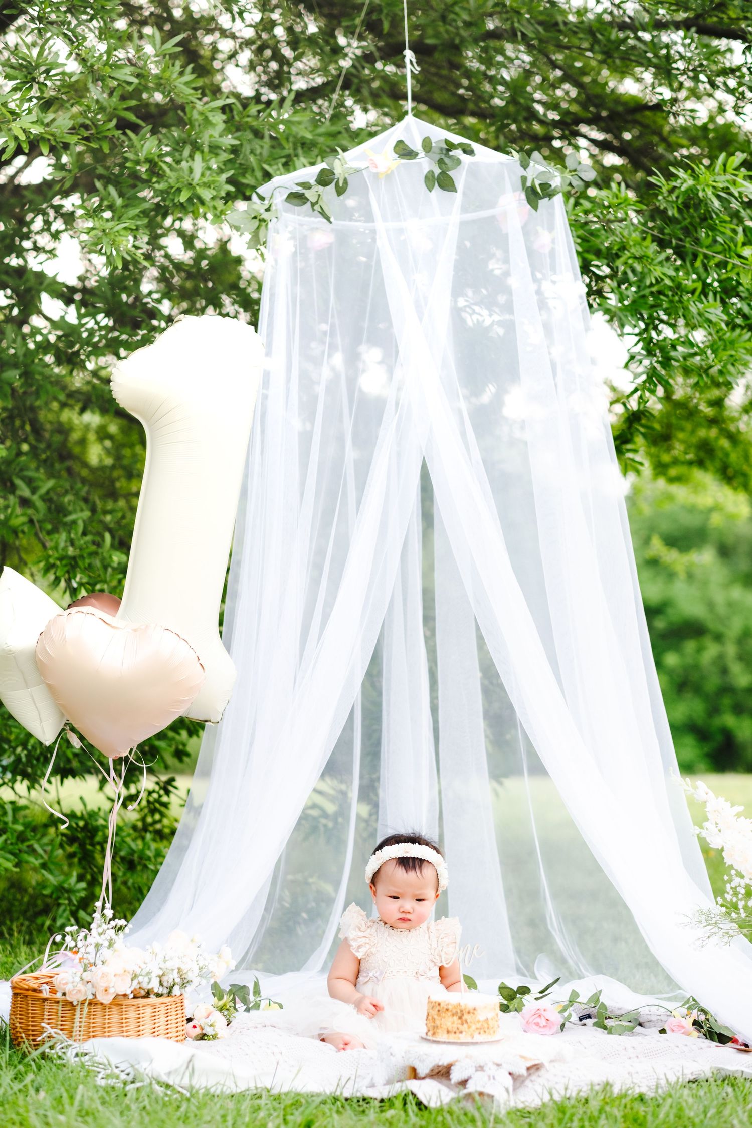 Baby girl in white dress sits under sheer canopy with number one balloon and cake for first birthday outdoor celebration.