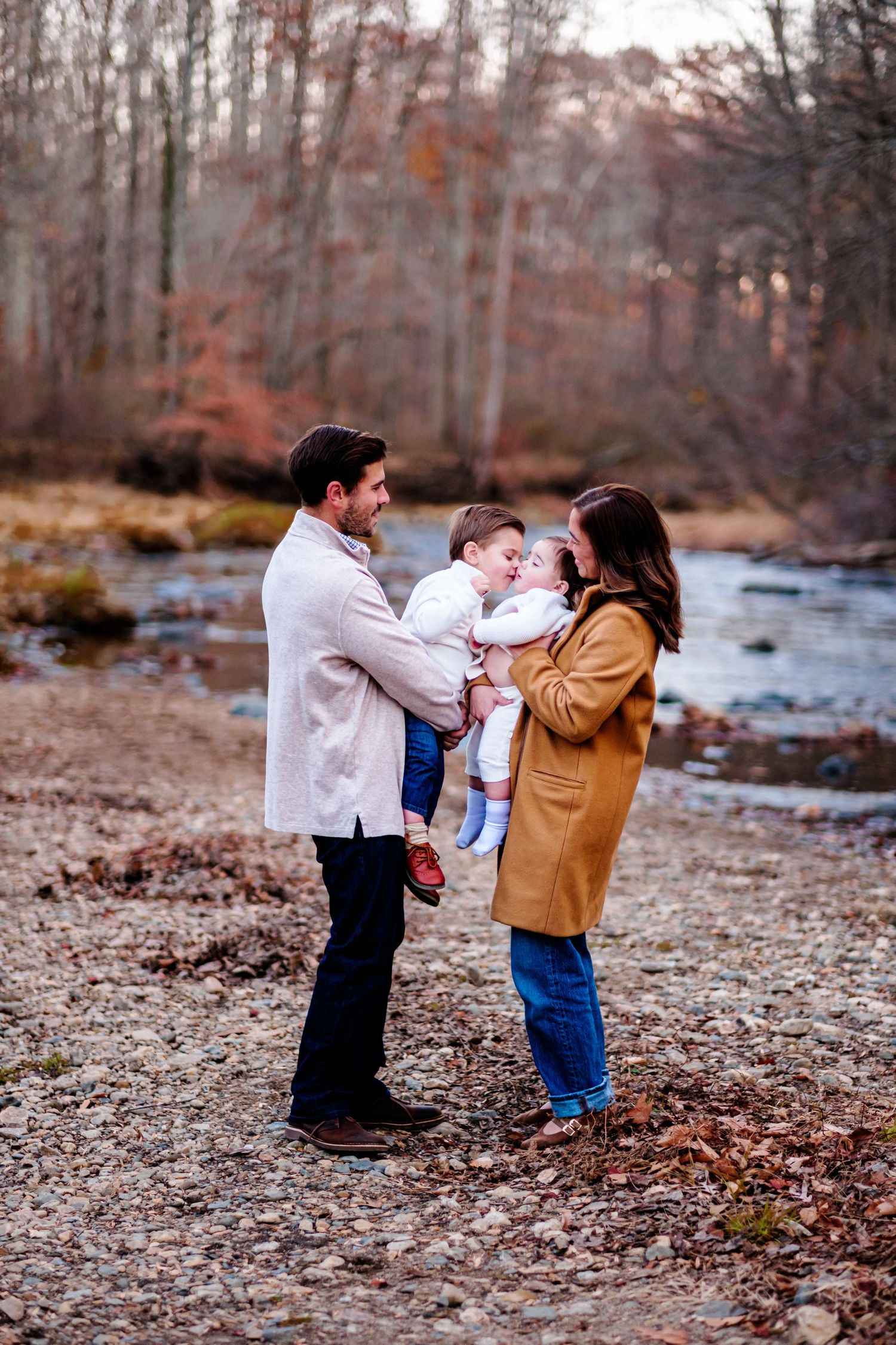 A couple holds their baby by a creek in autumn woods, wearing a white coat, tan coat, and jeans.