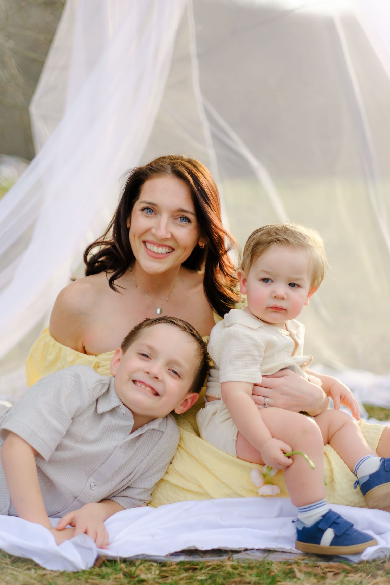 A smiling mother in a yellow dress sits outdoors with her two young sons under a white canopy on a sunny day.