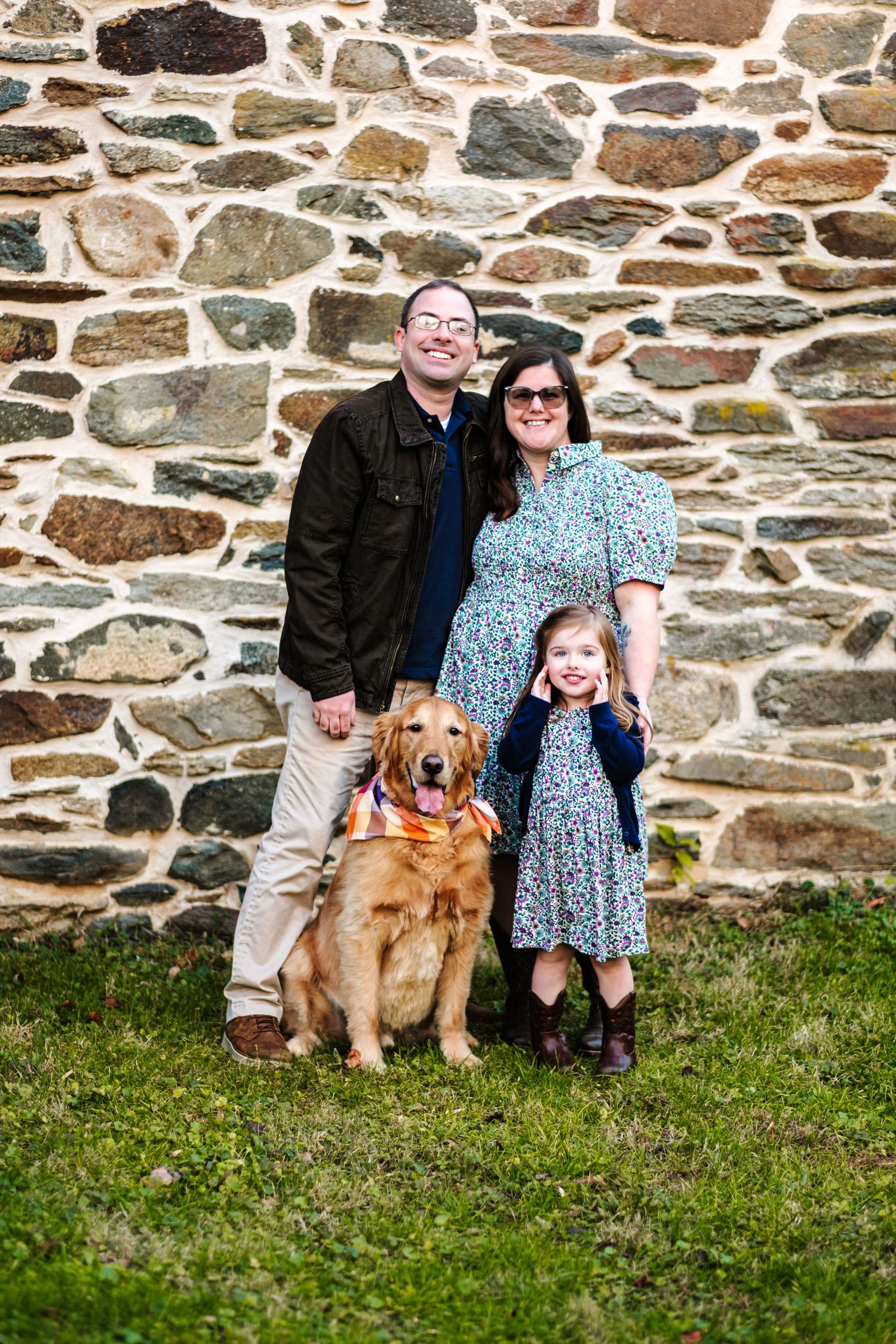 Family of two adults and a child posing with a golden retriever dog in front of a stone wall on green grass.