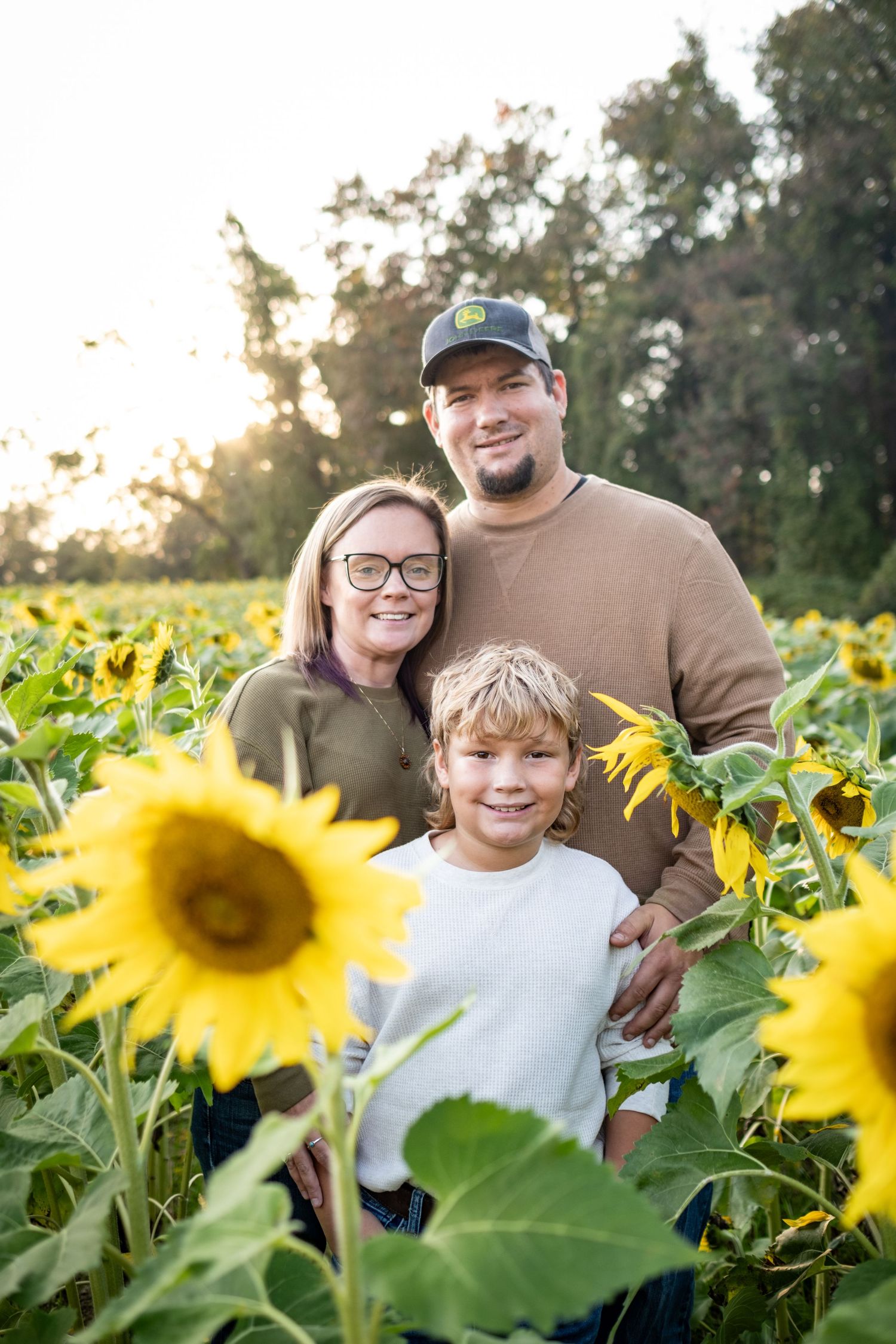 Family of three smiling in a sunflower field at golden hour, parents standing behind young boy surrounded by bright yellow blooms.