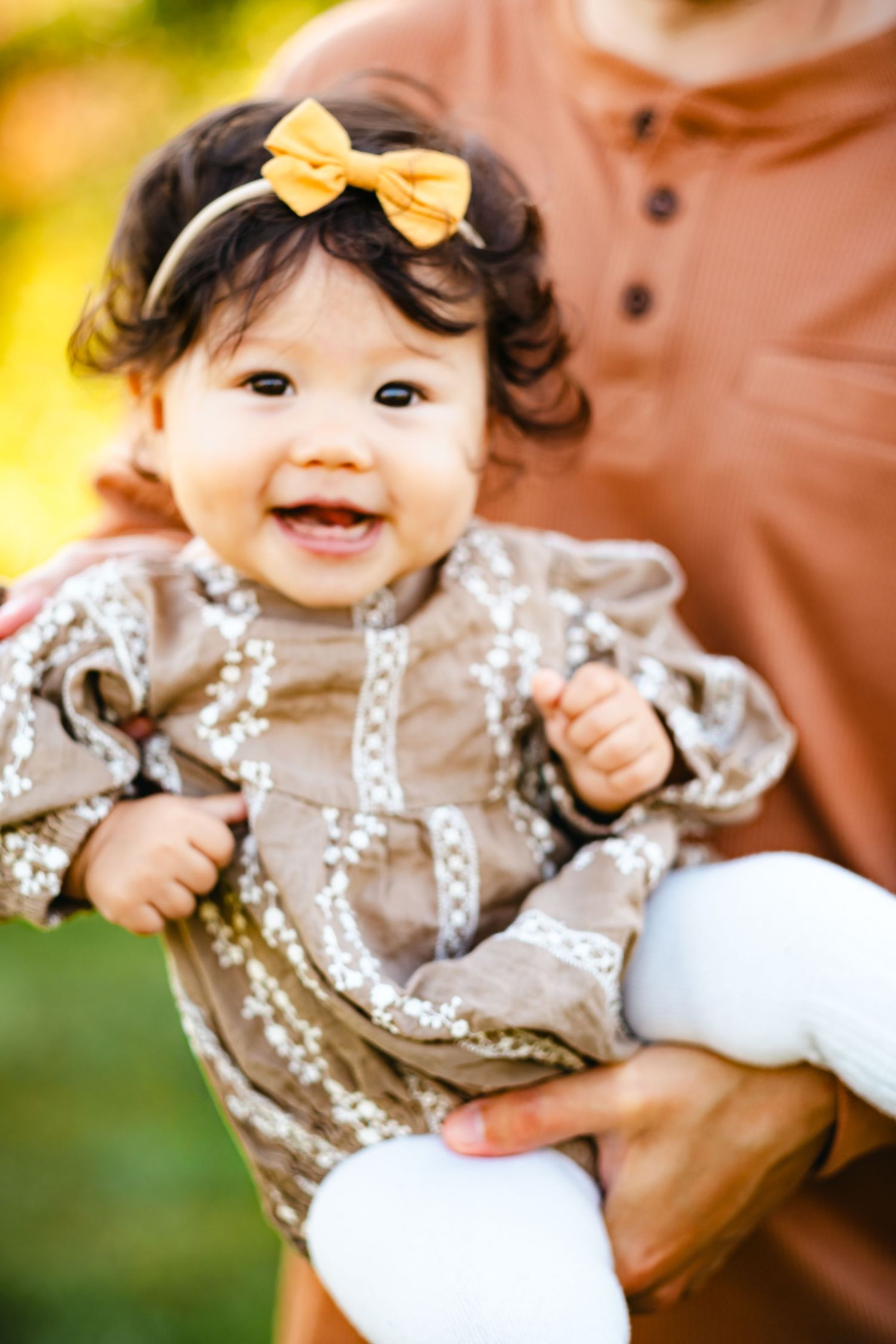 Smiling baby girl in tan embroidered dress and yellow bow headband being held outdoors.