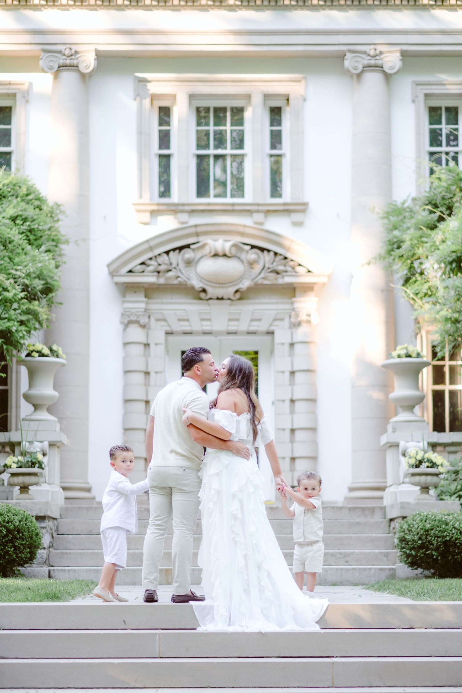 A couple in wedding attire kisses on mansion steps while two young children in white stand beside them.