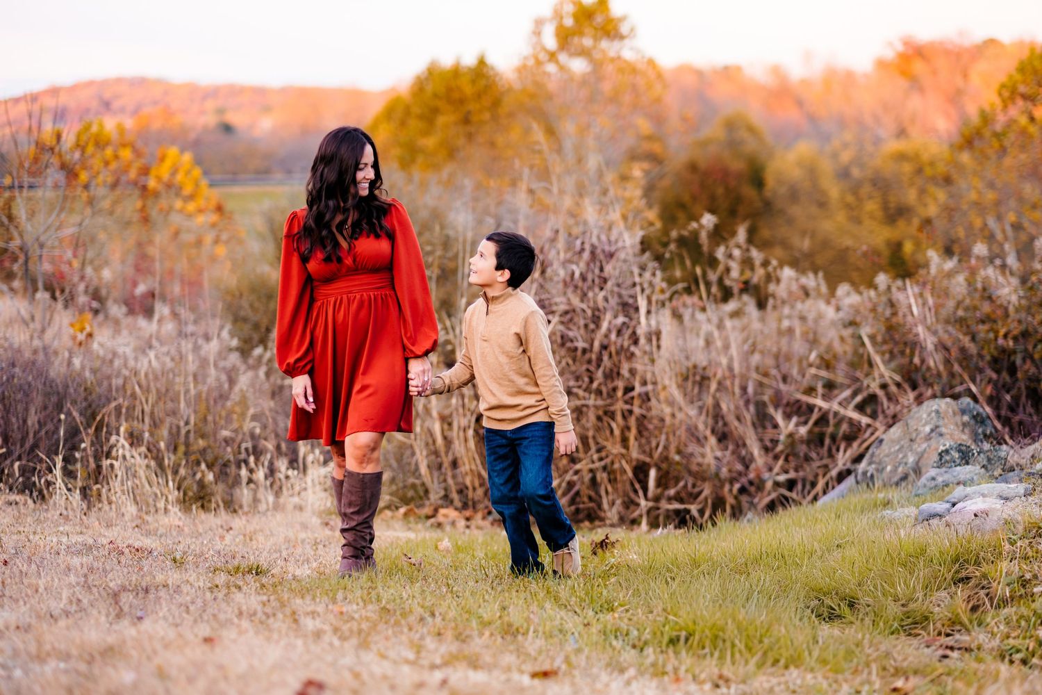 A mother in a red dress and brown boots holds hands with her young son in an autumn outdoor setting.