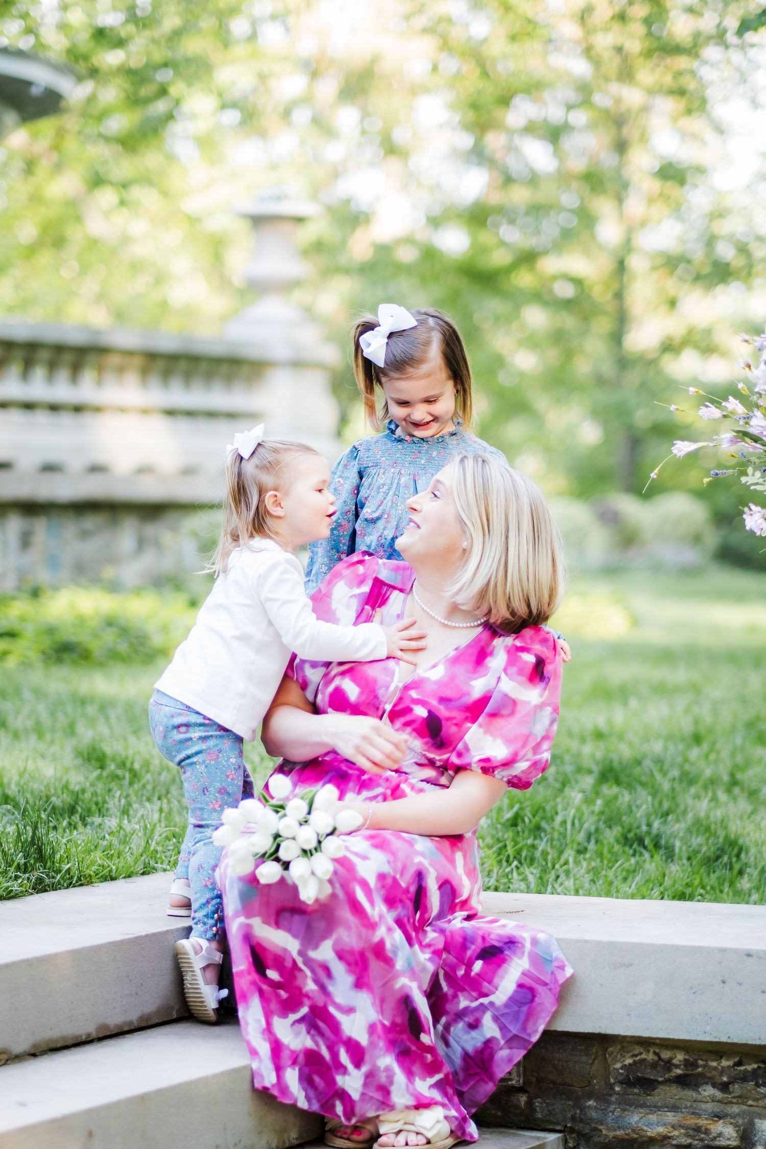 Pregnant woman in pink dress sits outdoors with two young daughters, sharing a tender moment in a lush green park.