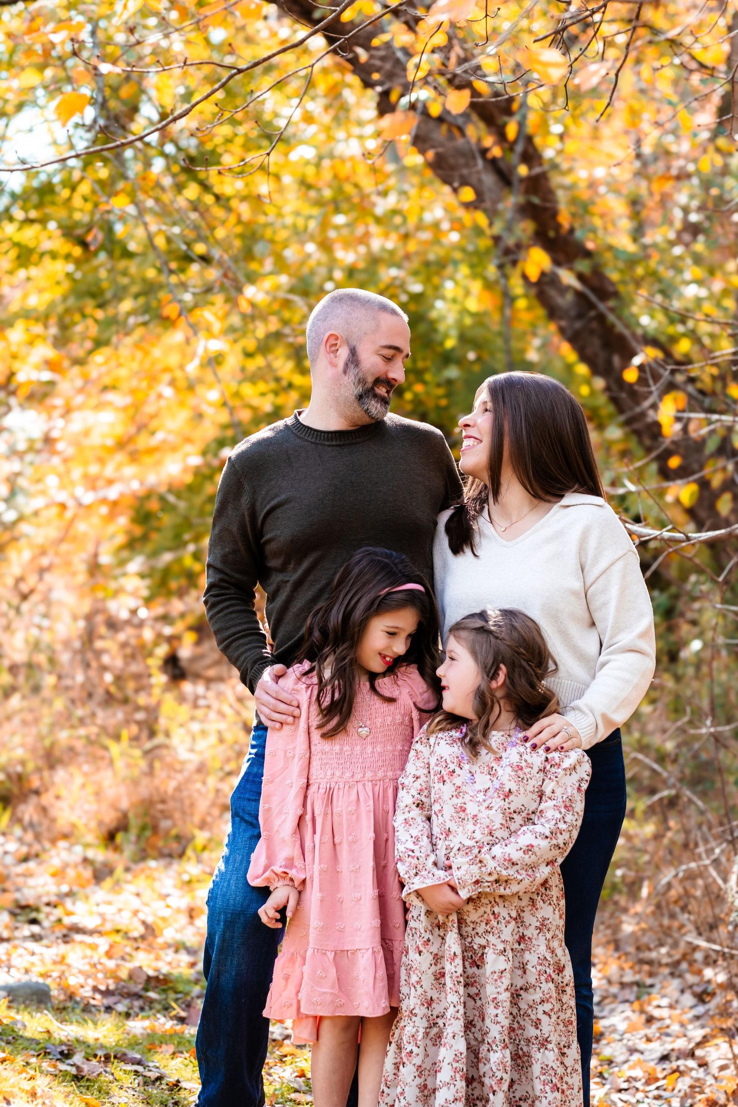 Family of four poses outdoors among golden autumn leaves, parents smiling at each other while hugging their two young daughters.