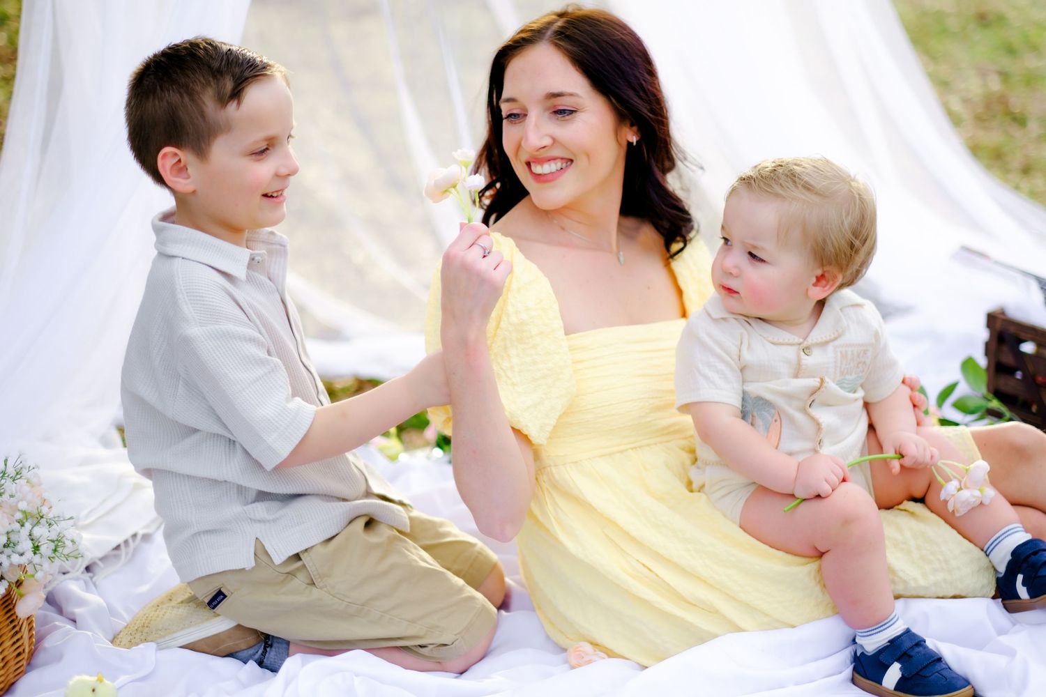 A smiling mother in a yellow dress sits outdoors with her two young sons during a sunny picnic setting.
