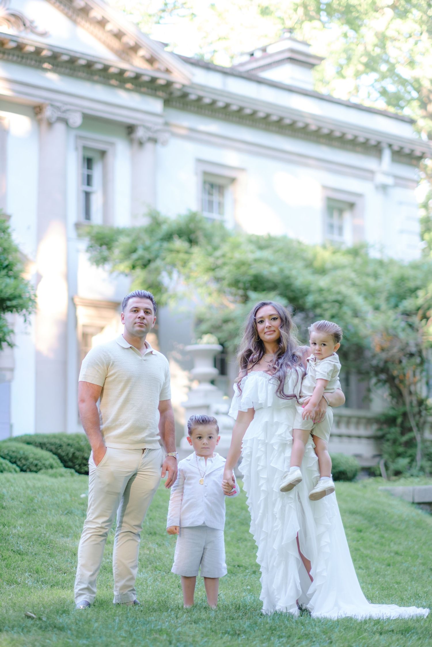 Family of four posing outdoors in elegant attire before a grand estate, with lush greenery and classical architecture.