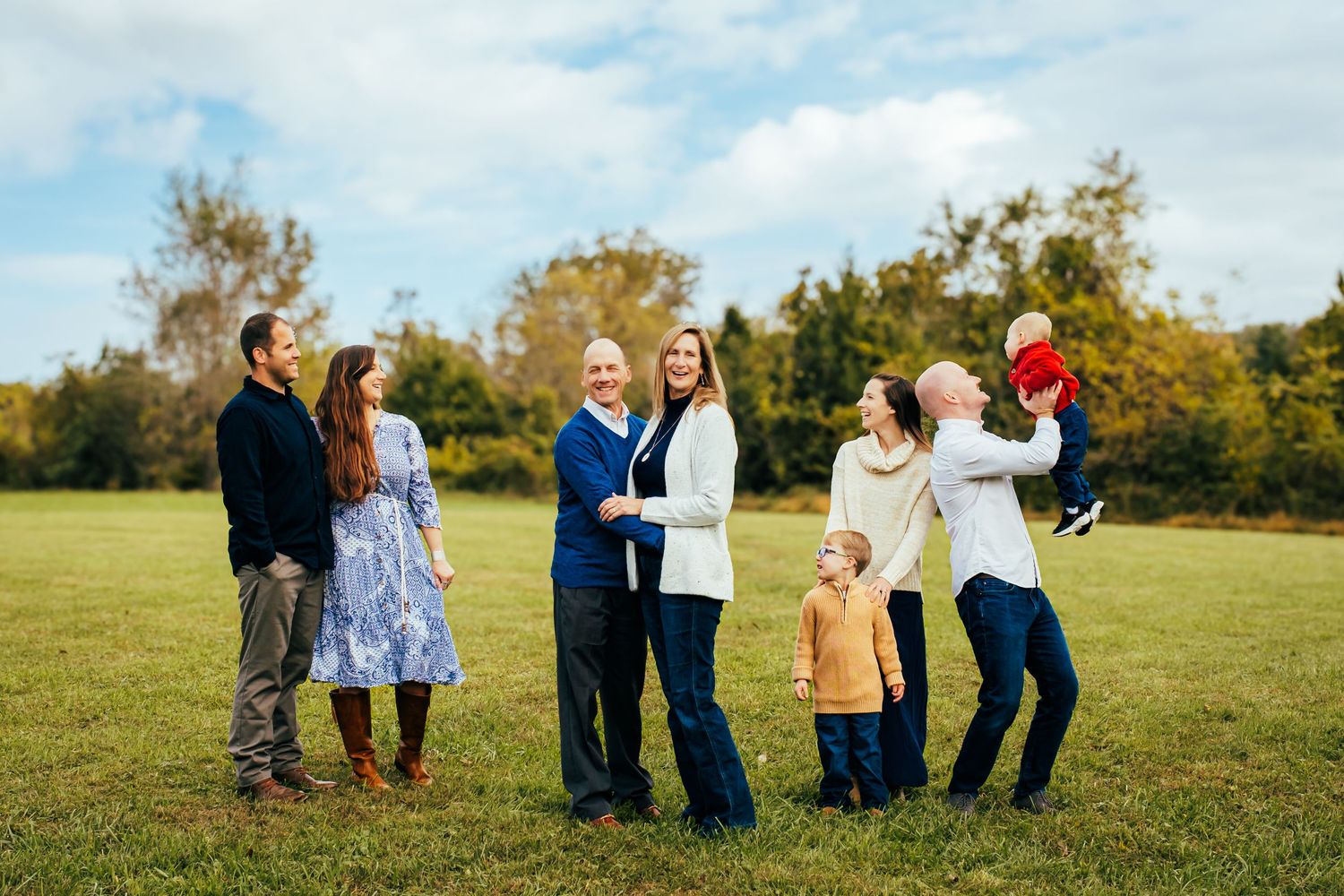 Multi-generational family posing outdoors in a green park, smiling and playing together on a sunny day with trees in background.