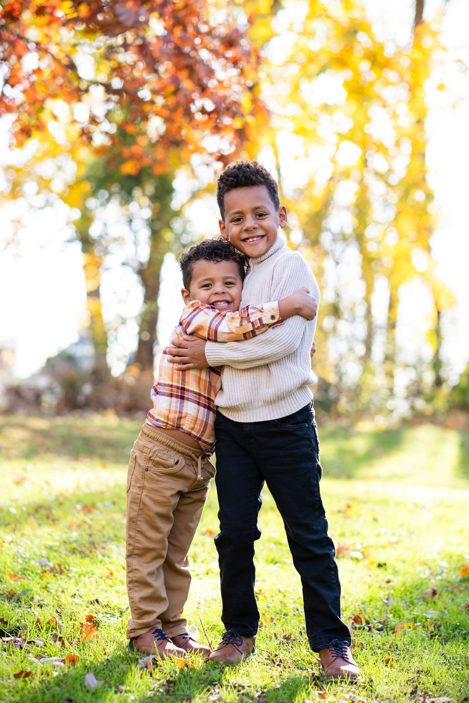 Two young boys hugging and smiling outdoors during autumn, surrounded by colorful fall foliage.